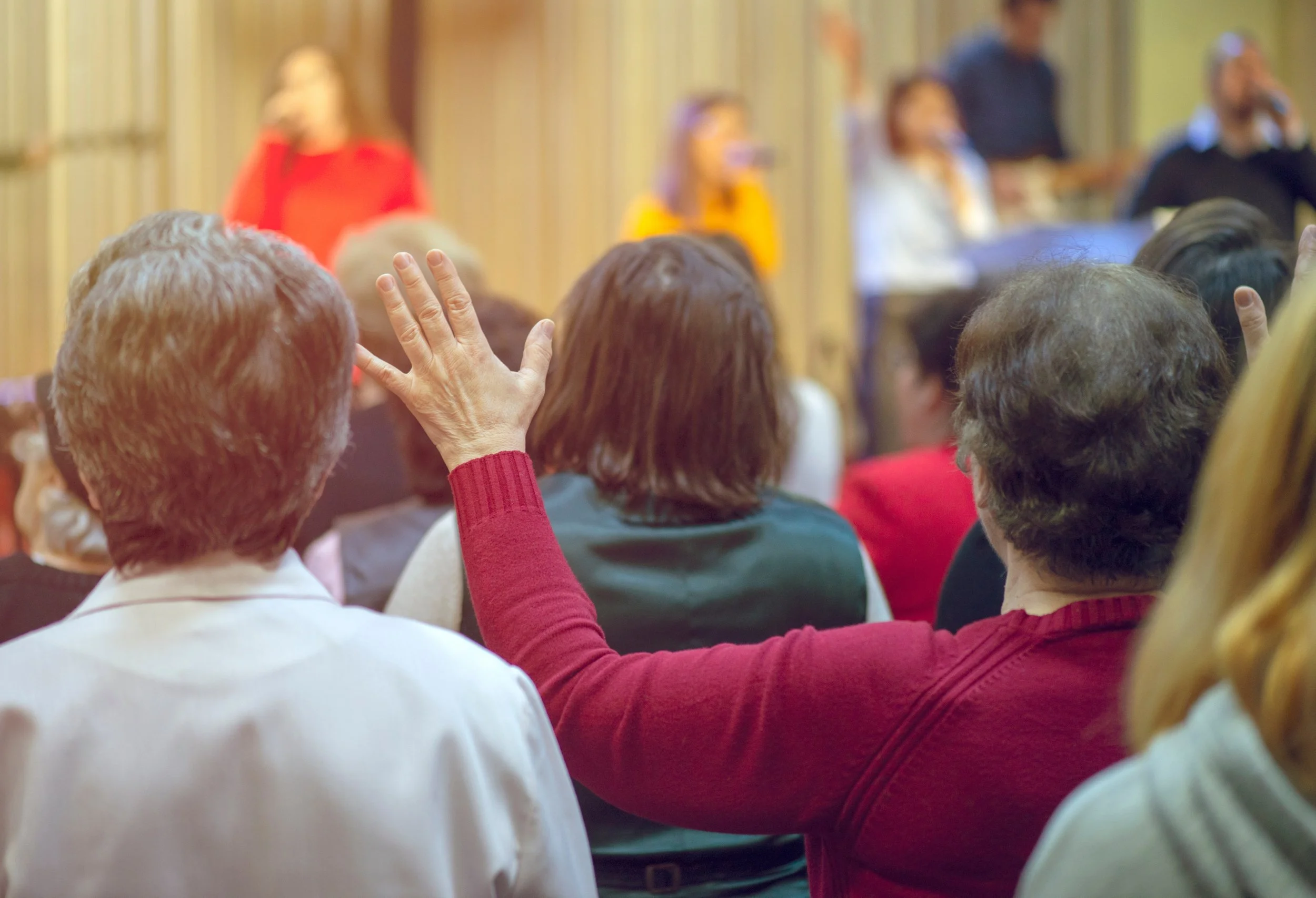 People attending a seminar or conference, with one person raising their hand to ask a question, in a room with wood-paneled walls.