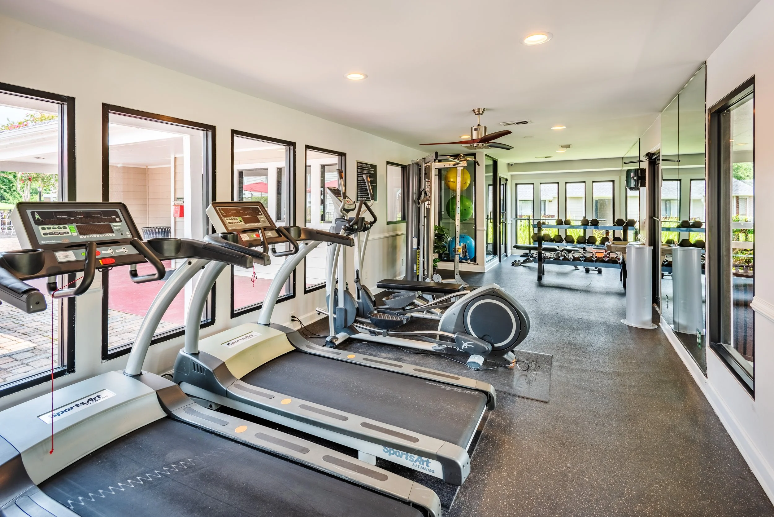Empty indoor gym with treadmills, elliptical trainer, weight rack, stability balls, and windows.