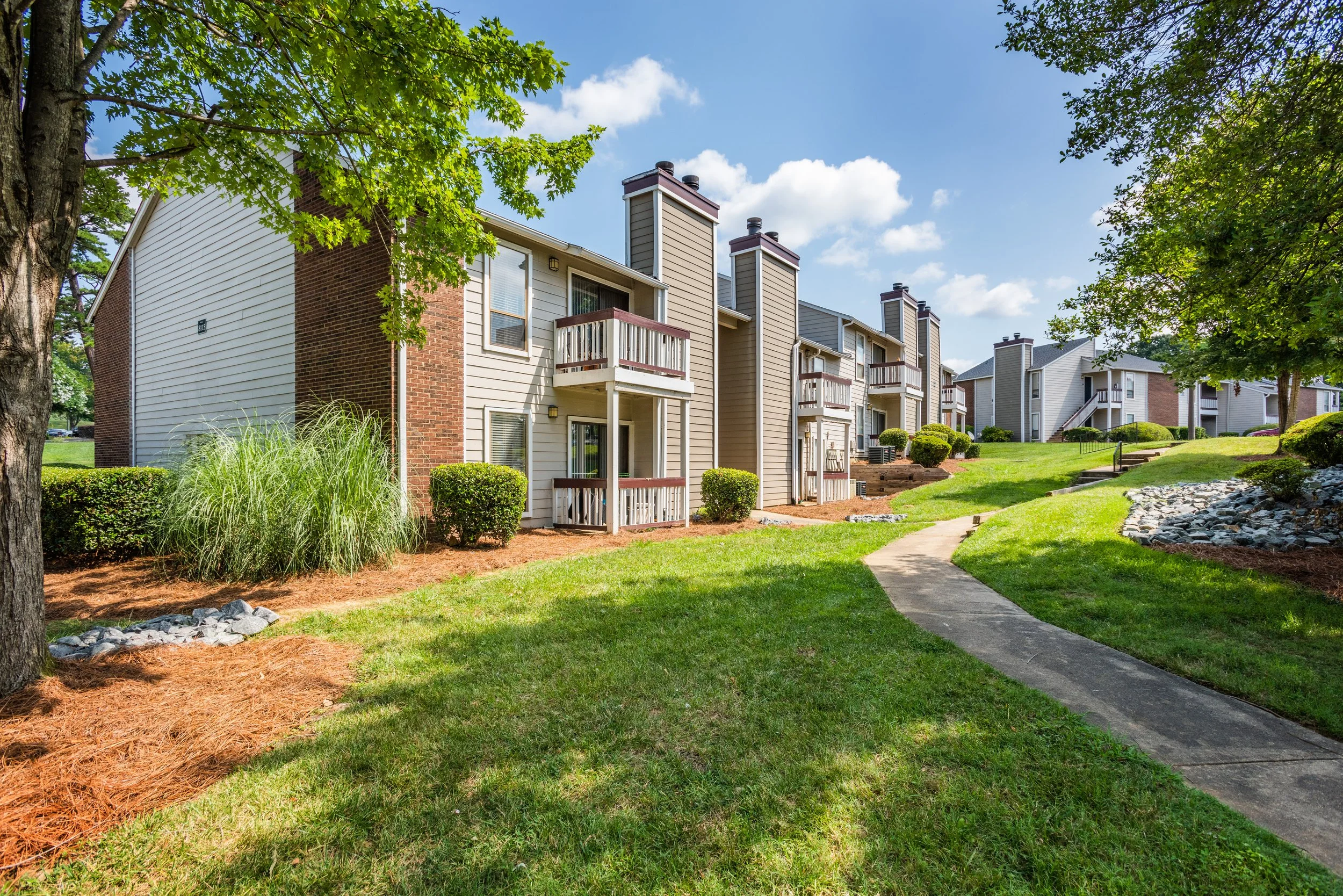 Multi-unit residential apartment complex with grassy lawns, trees, shrubs, and a curved concrete walkway on a sunny day.