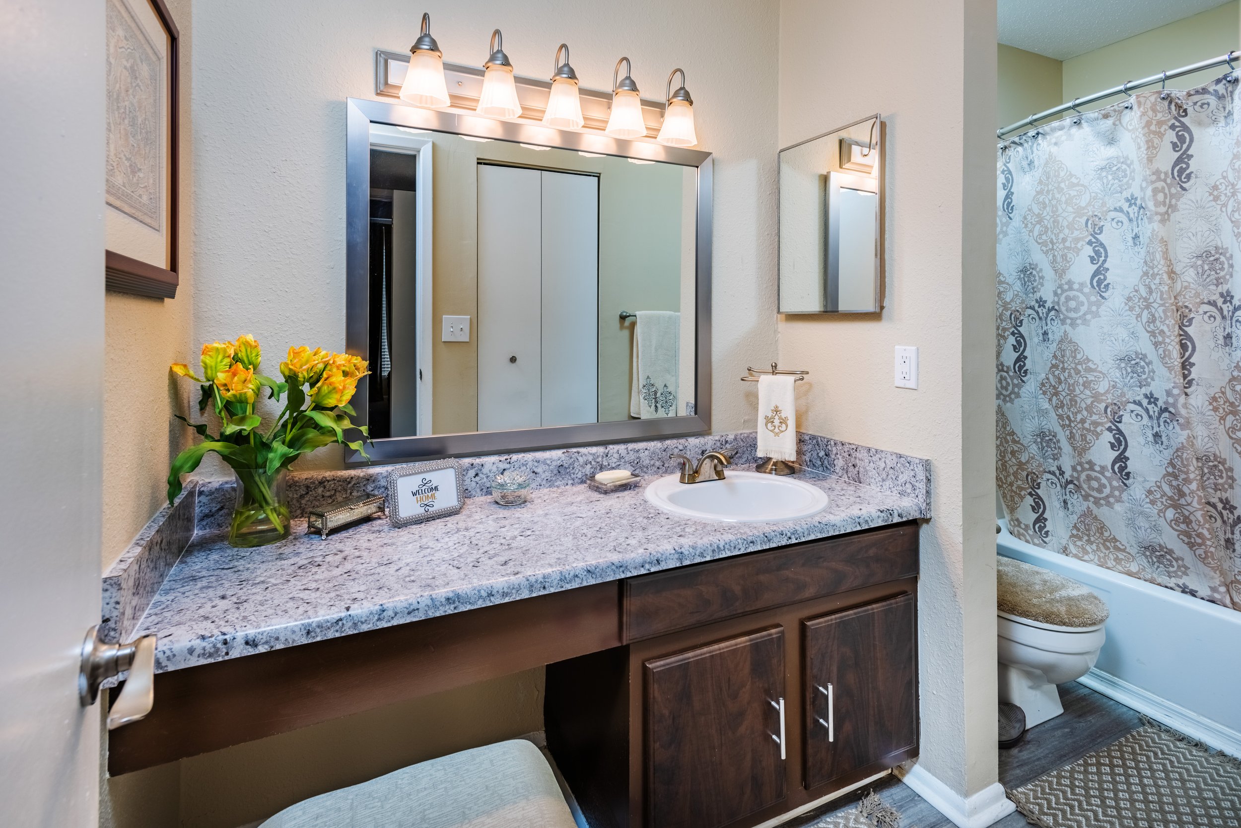 Bathroom vanity with a granite countertop, mirror, light fixture, decorative items, a towel, and a flower arrangement, with a toilet and shower curtain visible in the background.