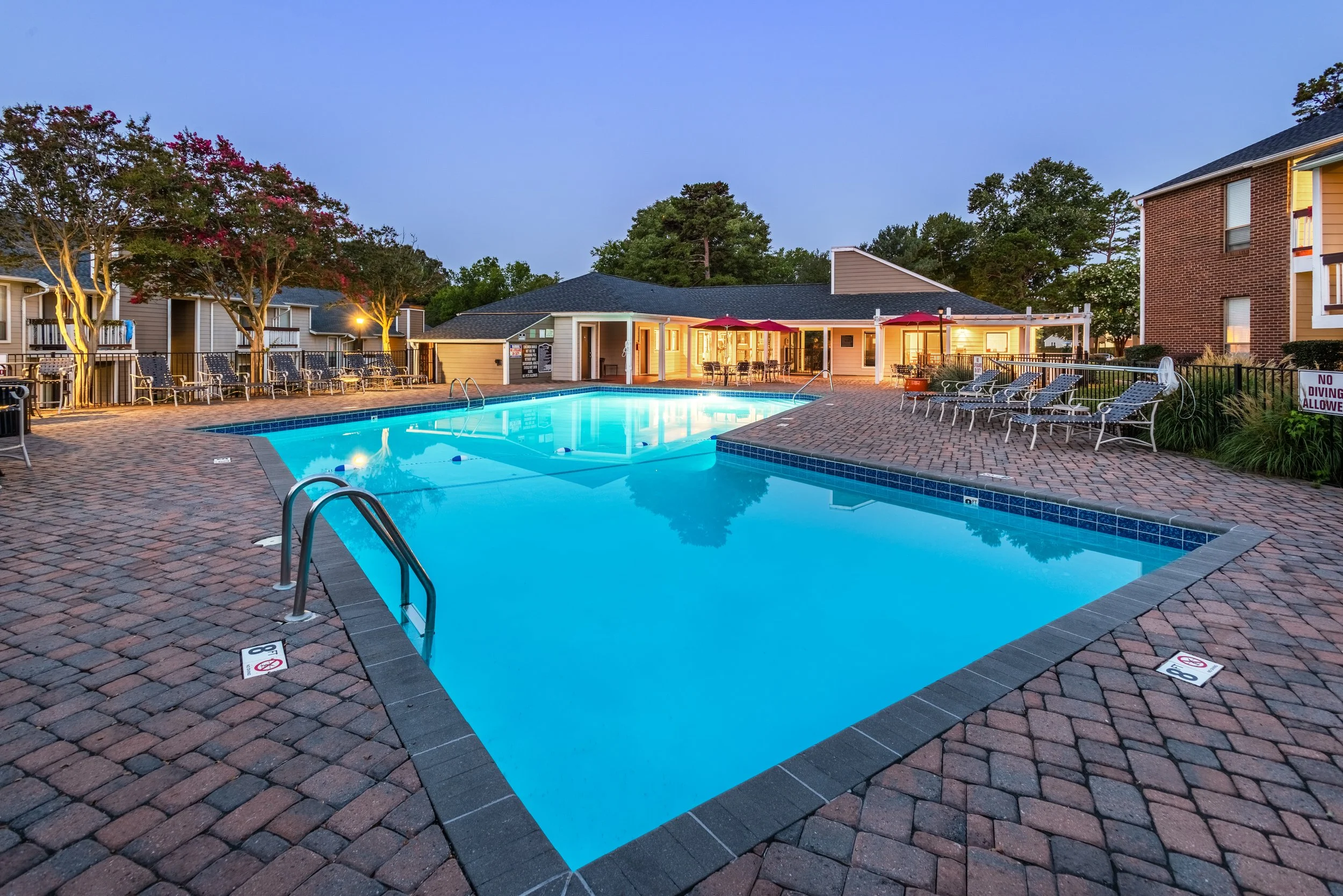 An outdoor community swimming pool at dusk, surrounded by lounge chairs, trees, and residential buildings, with a clubhouse in the background.