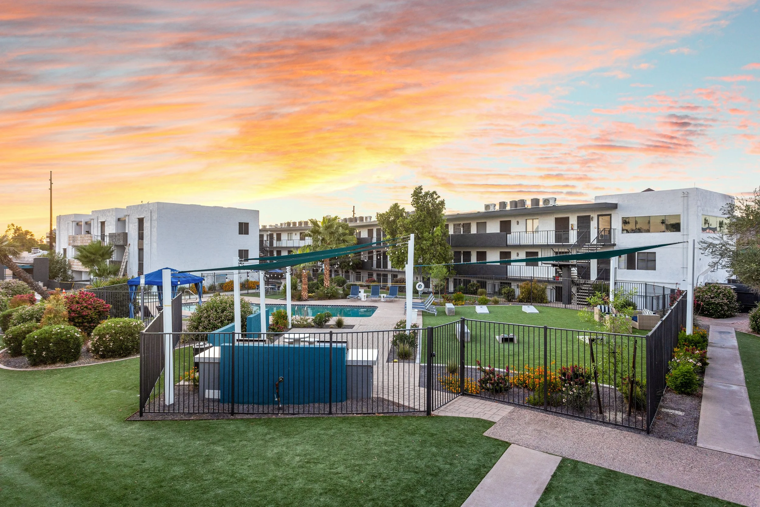 An apartment complex courtyard at sunset with a swimming pool, lounge chairs, greenery, and a gated area, under a colorful sky with orange and pink clouds.