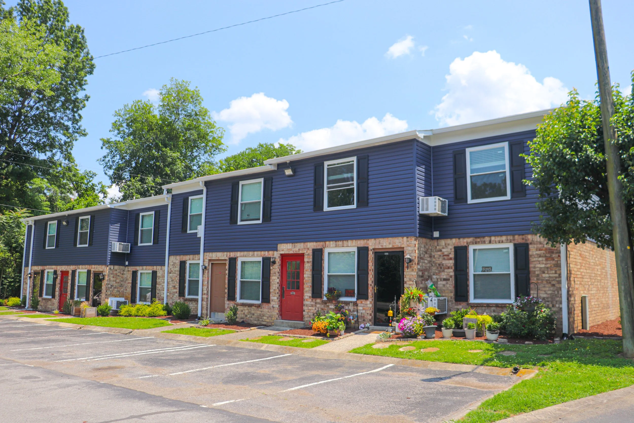 Multi-unit residential building with blue siding, brick lower level, black shutters, and red doors, surrounded by a parking lot and landscaped with bushes and flowers, under a partly cloudy sky.