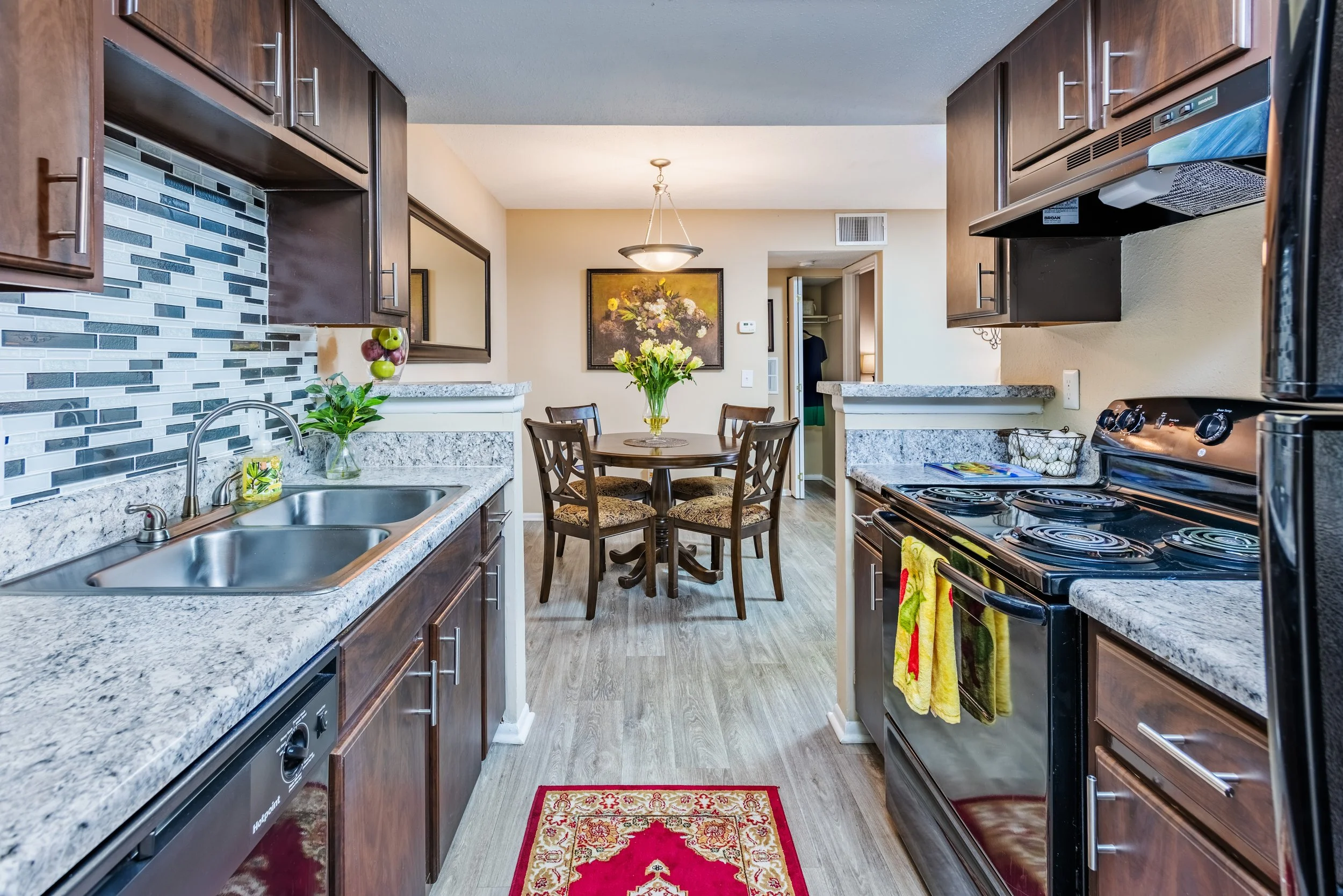 View from kitchen looking into dining area with round table, four chairs, and a vase of flowers. Kitchen has dark wood cabinets, granite countertops, a double sink, backsplash with gray and black tiles, and appliances including a stove and refrigerat