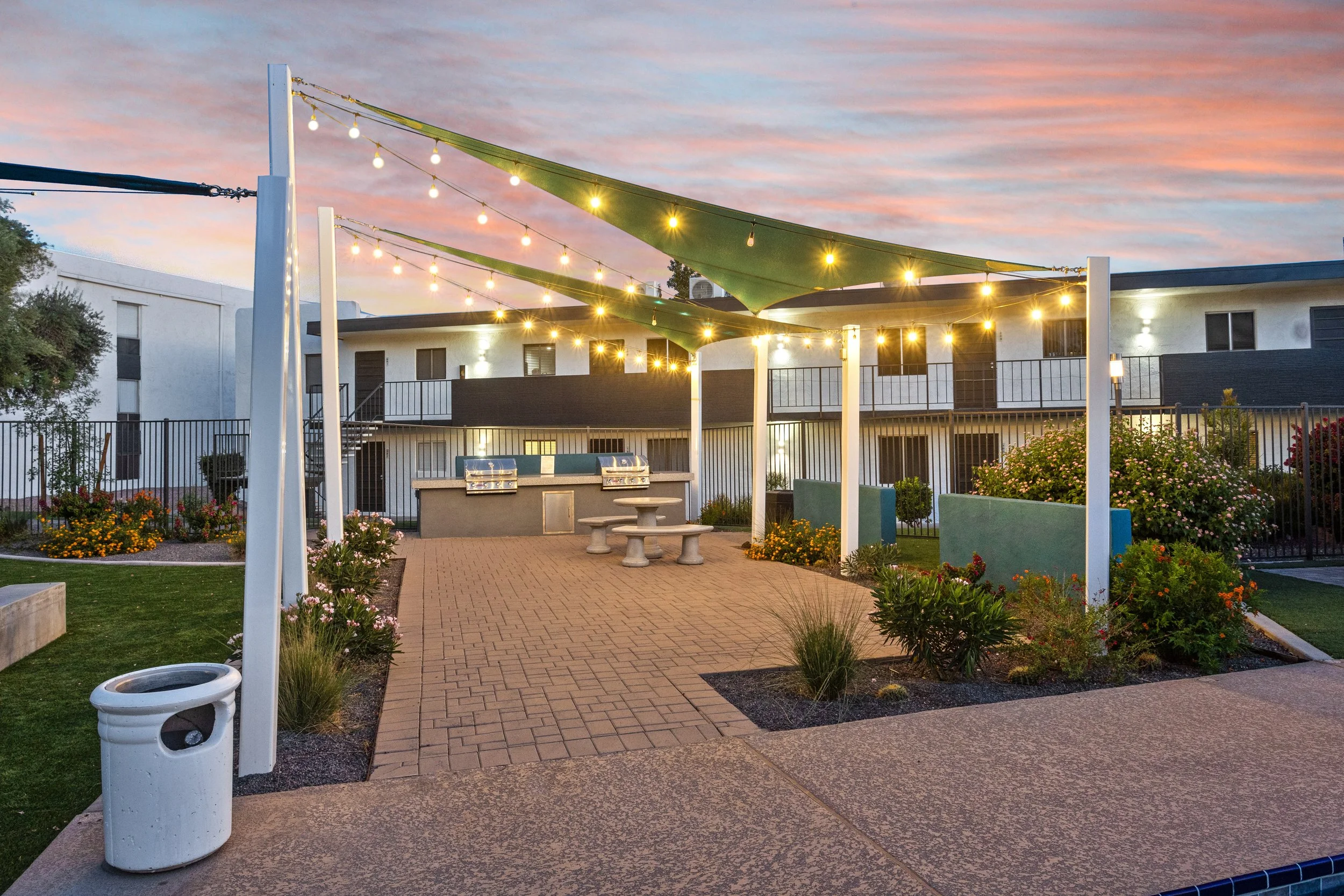 Outdoor courtyard with string lights and fabric shade sails over a paved area, featuring a barbecue grill and seating, surrounded by garden beds and apartment buildings at dusk.