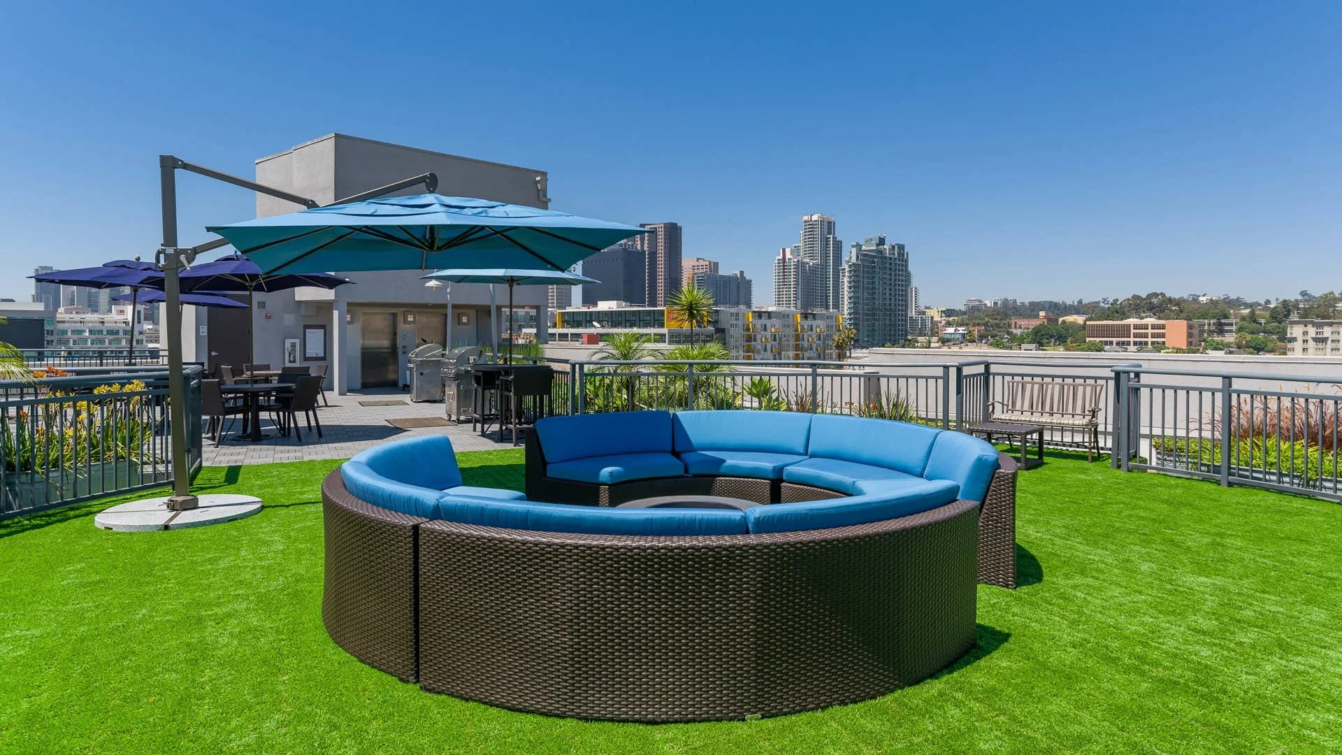 Rooftop outdoor lounge with a circular sectional sofa with blue cushions, green artificial grass, umbrellas, tables, and a city skyline in the background under clear blue sky.