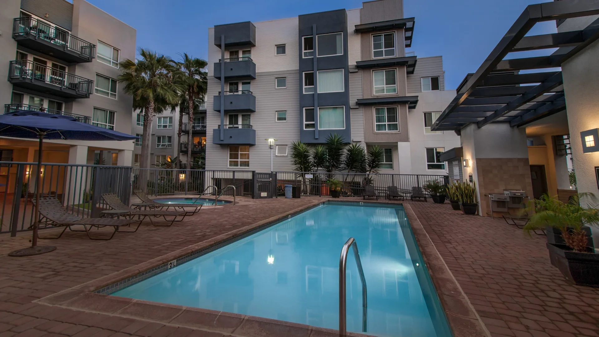 A swimming pool area in a modern apartment complex during dusk, with lounge chairs, a small hot tub, potted plants, and high-rise residential buildings in the background.