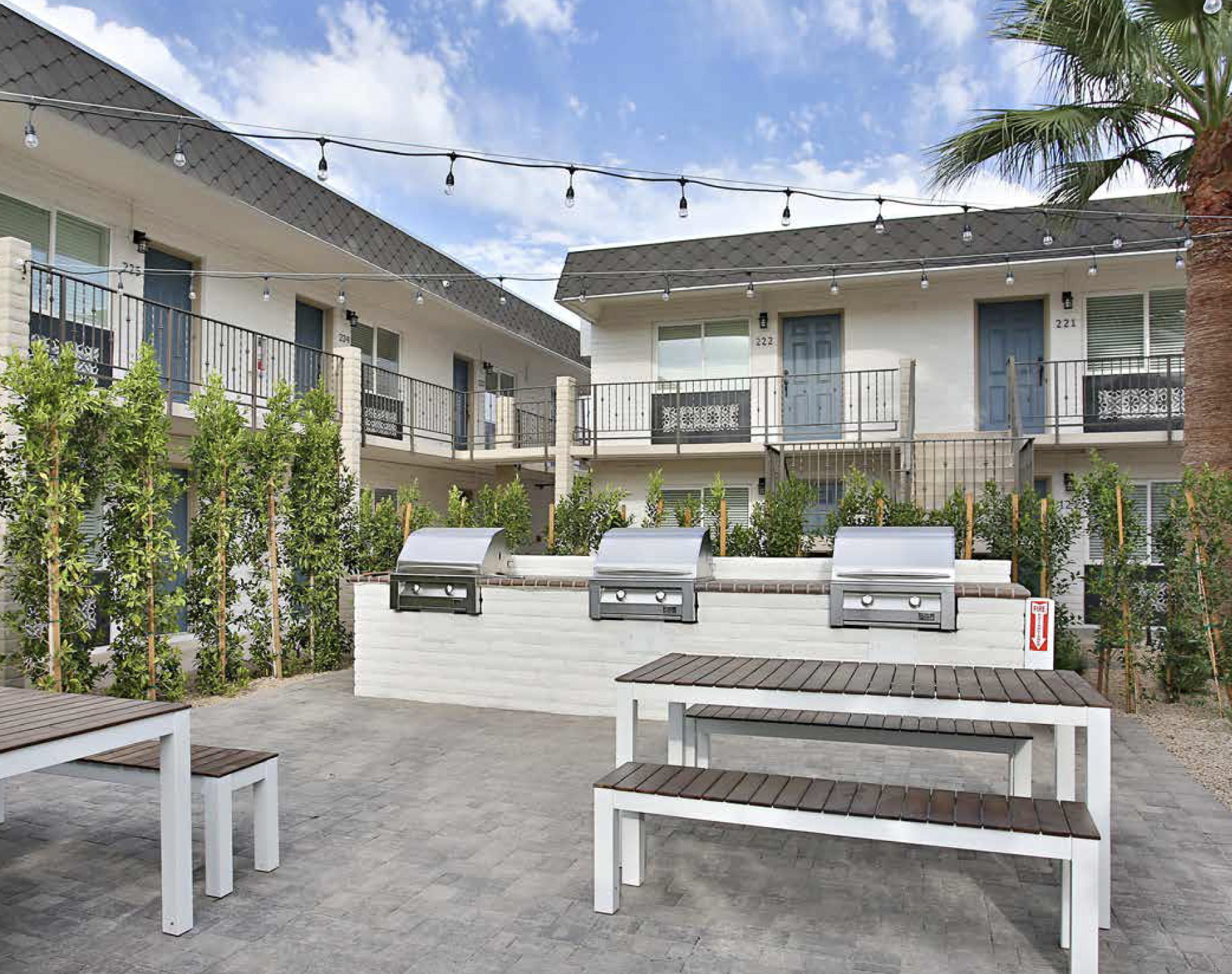 Outdoor communal space with stone-paved ground, two picnic tables with wooden tops and white legs, three stainless steel gas grills, surrounded by greenery, apartment buildings with balconies and blue doors, string lights overhead, and a palm tree on