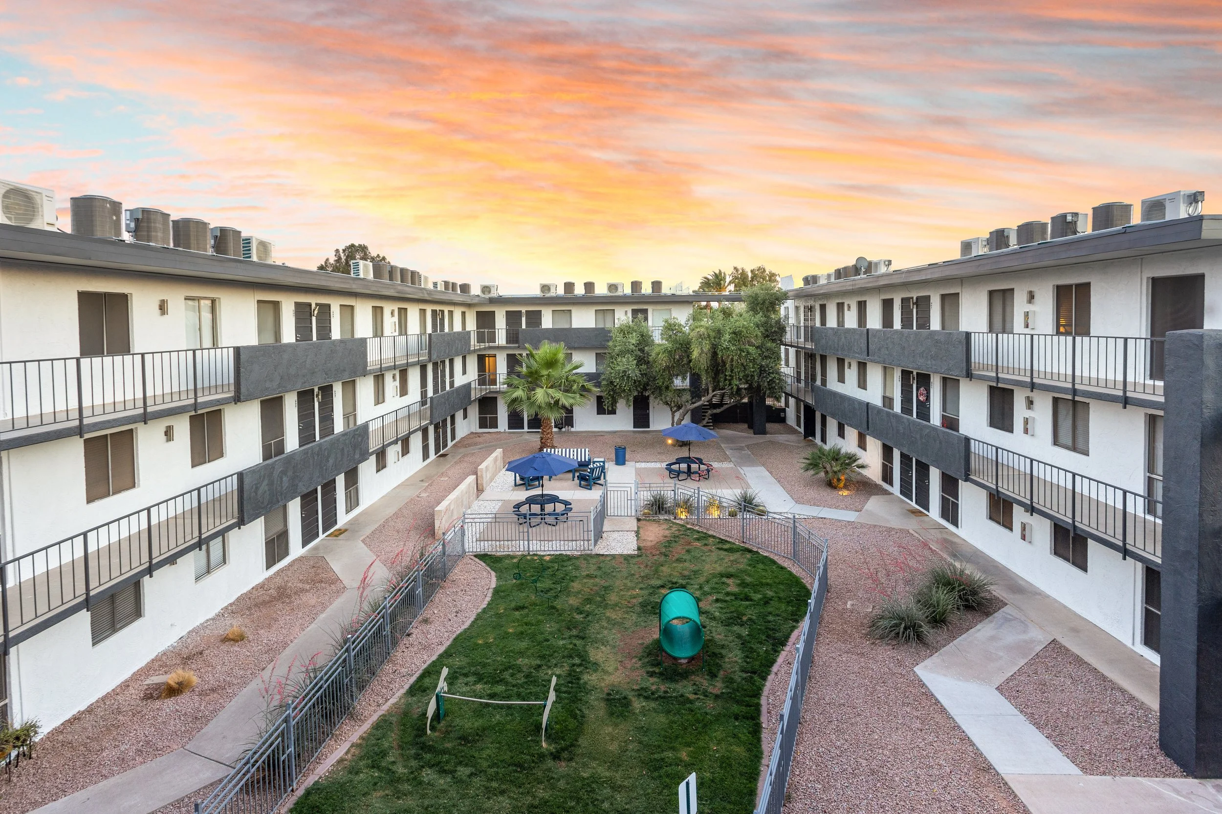Courtyard view of a mid-rise apartment complex with two-story buildings on each side, a grassy area with a small tunnel and benches in the center, surrounded by a metal fence, with trees, outdoor seating with umbrellas, and a colorful sunset sky in t