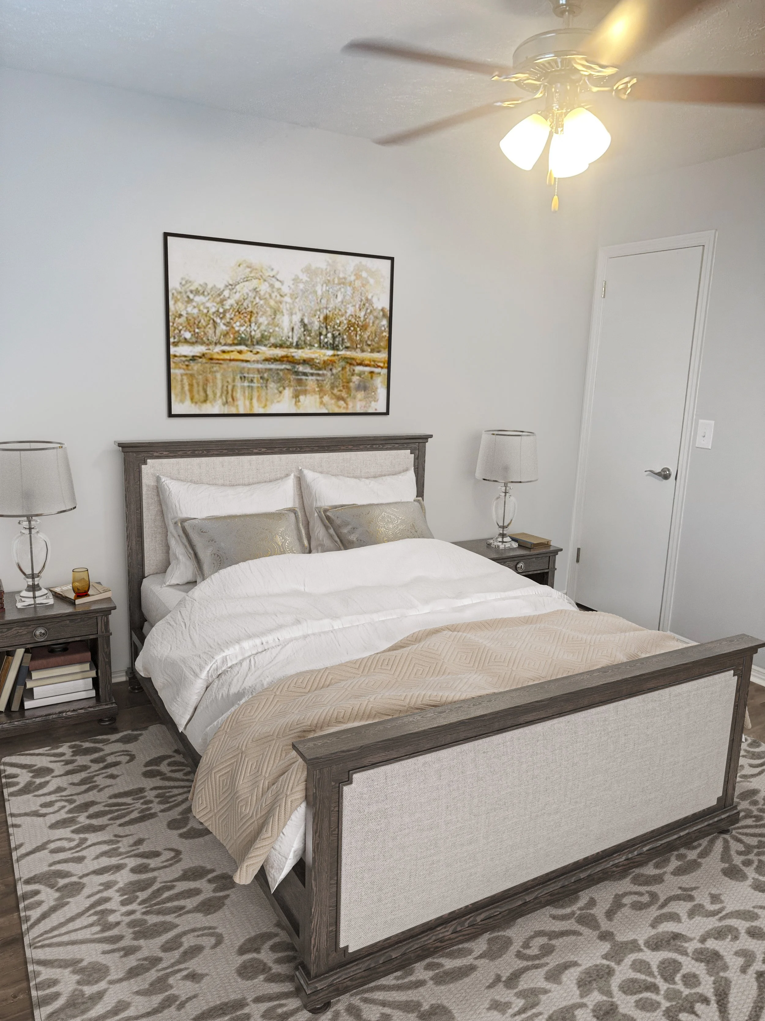 A bedroom with a large bed, gray headboard and footboard, white bedding, beige quilt, and decorative pillows. There are matching gray nightstands with lamps and books. A landscape painting hangs above the bed. The room has a ceiling fan with lights, 
