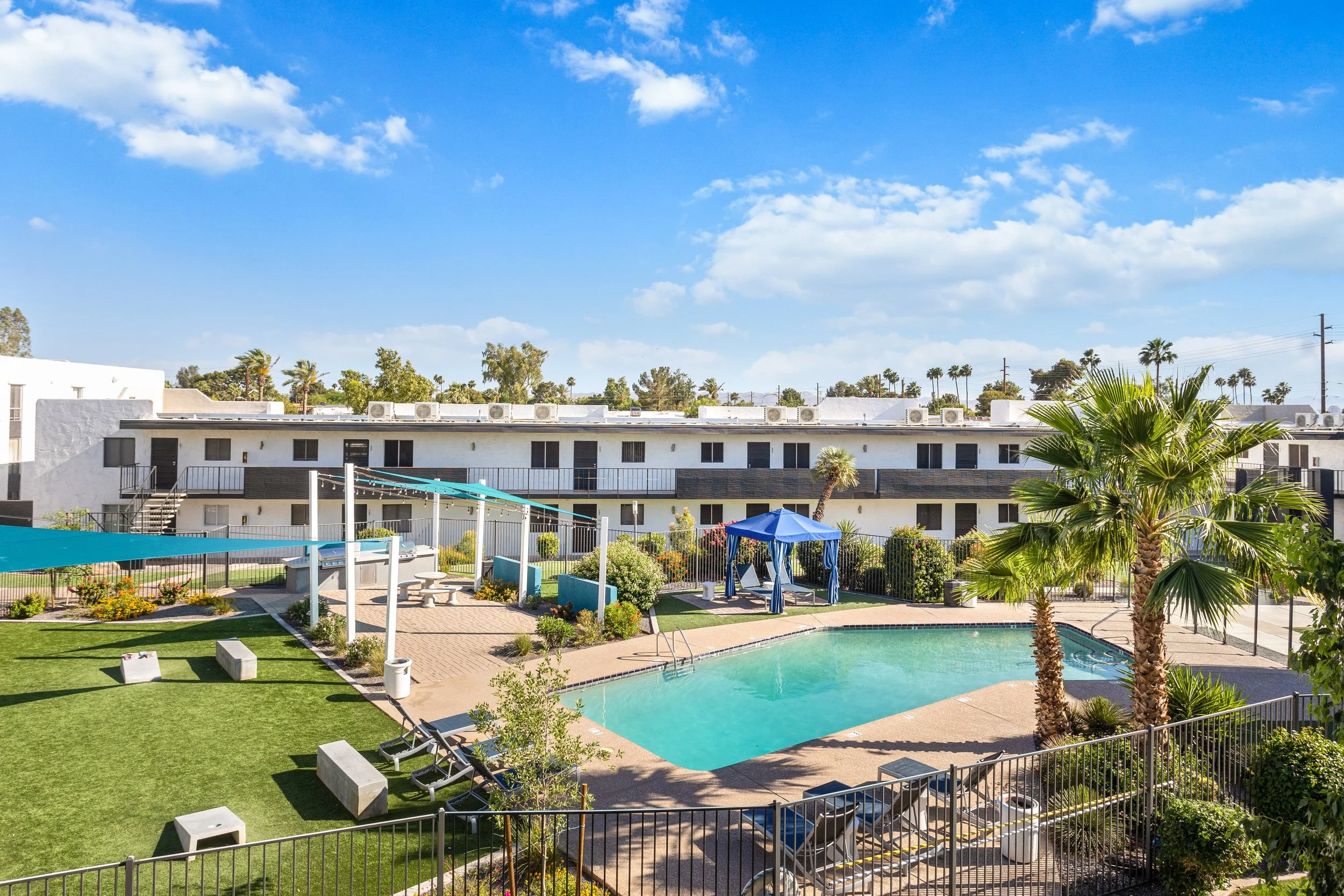 A residential apartment complex with a swimming pool, palm trees, lounge chairs, and a shaded gazebo under a blue sky with scattered clouds.