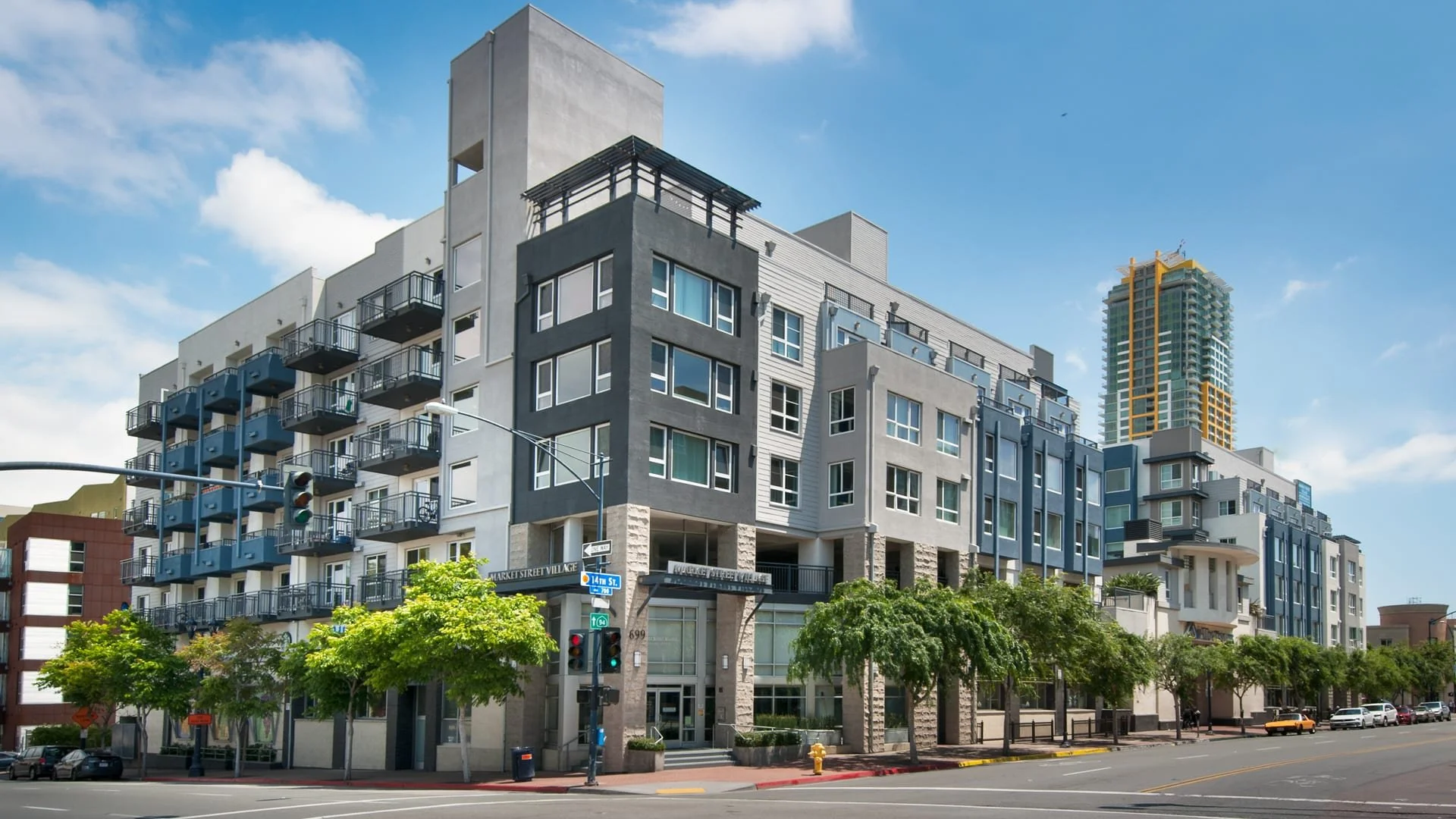 Modern multi-story apartment building with balconies, trees along the sidewalk, and a busy street corner with traffic lights.