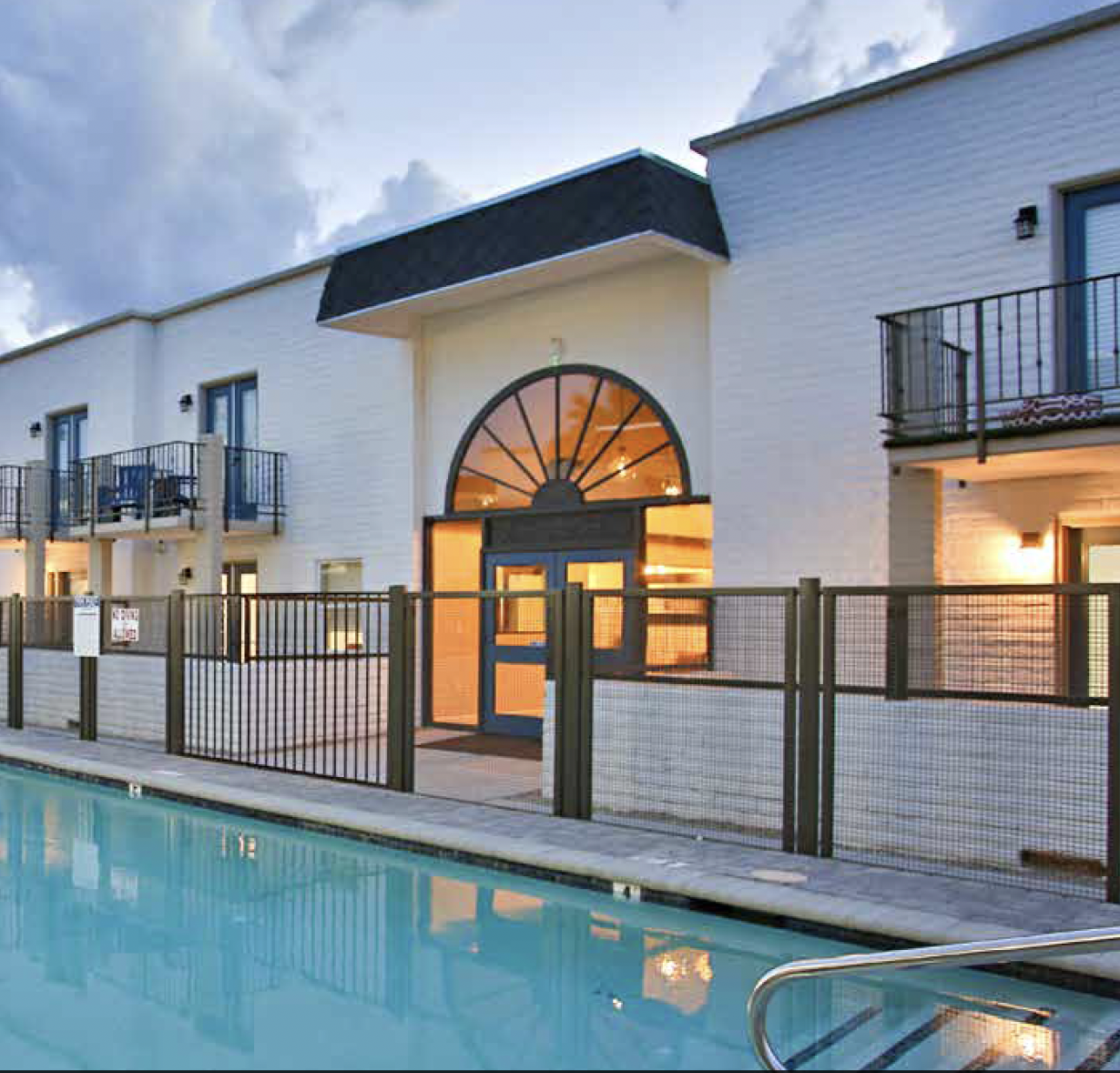 View of a residential apartment complex with a swimming pool in the foreground and a white building with balconies and a large arched window in the background, during dusk.