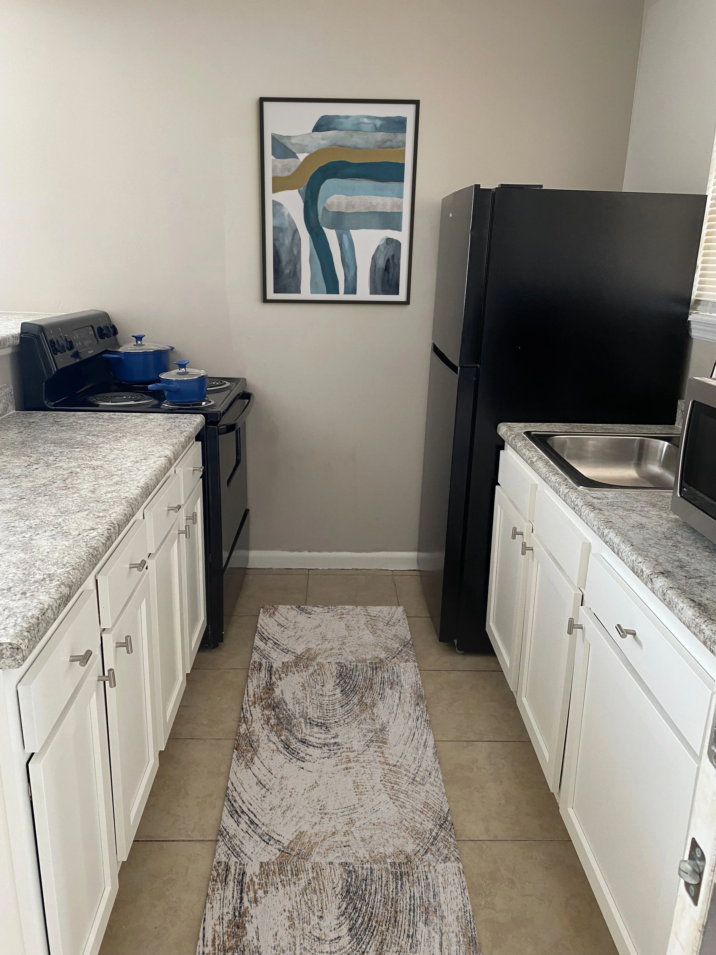 Small kitchen with white cabinets, a gray countertop, black refrigerator, microwave, blue pots on stove, and a framed abstract painting on the wall.