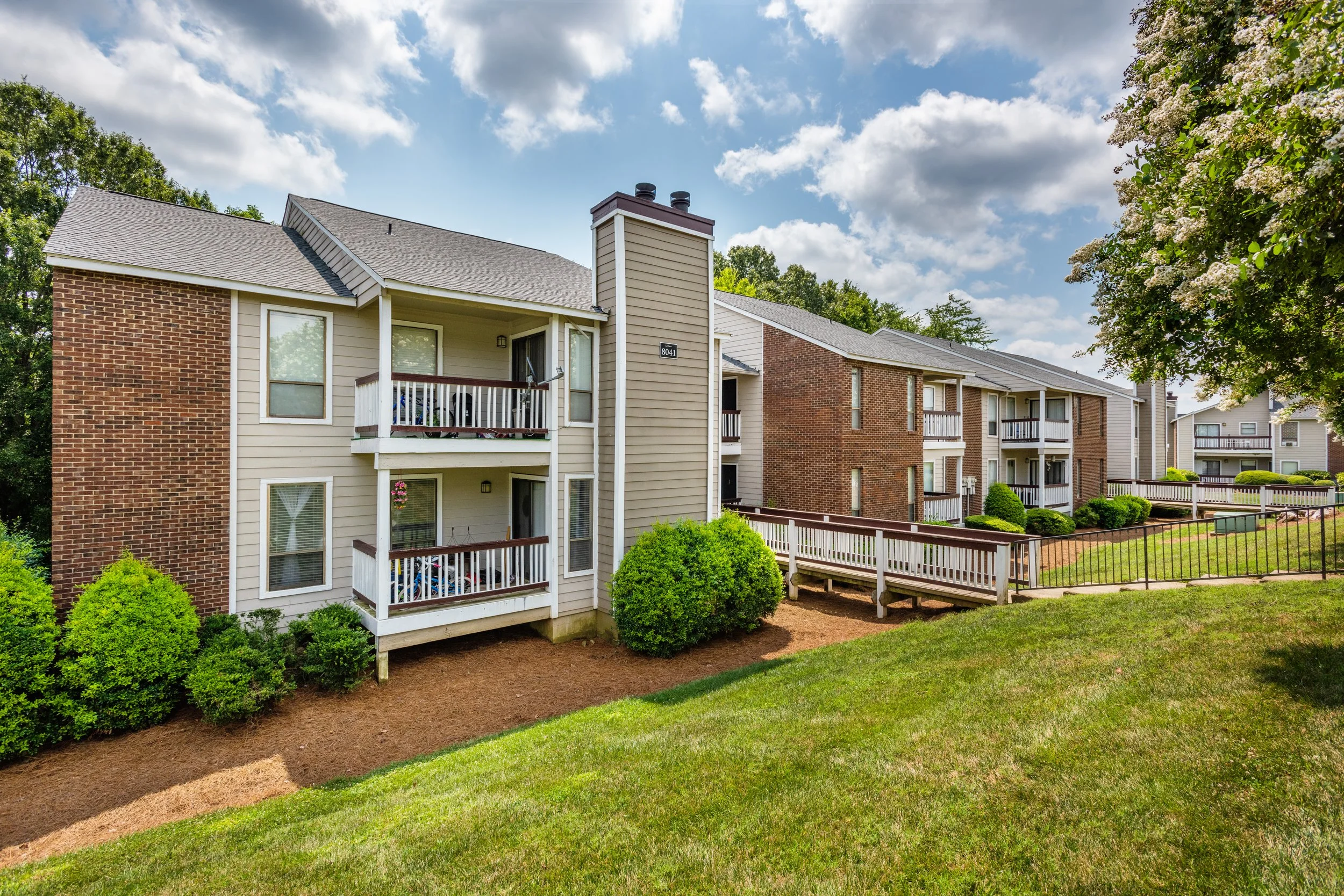 Multi-story residential apartment complex with beige and brick exterior, white balconies, well-manicured green lawns, shrubbery, and a wooden ramp, under a partly cloudy sky.