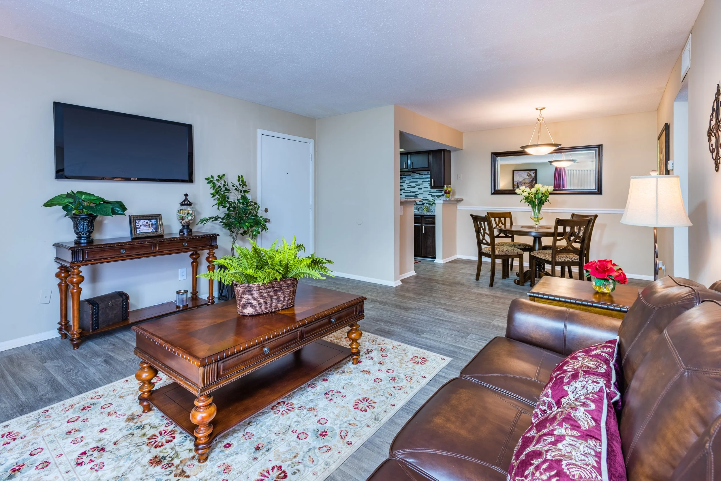 Living room with a leather sofa, wooden coffee table with plants, wall-mounted flat-screen TV, a side table with flowers, and a dining area with a round table and four chairs in the background. The room has wood-look flooring and neutral-colored wall
