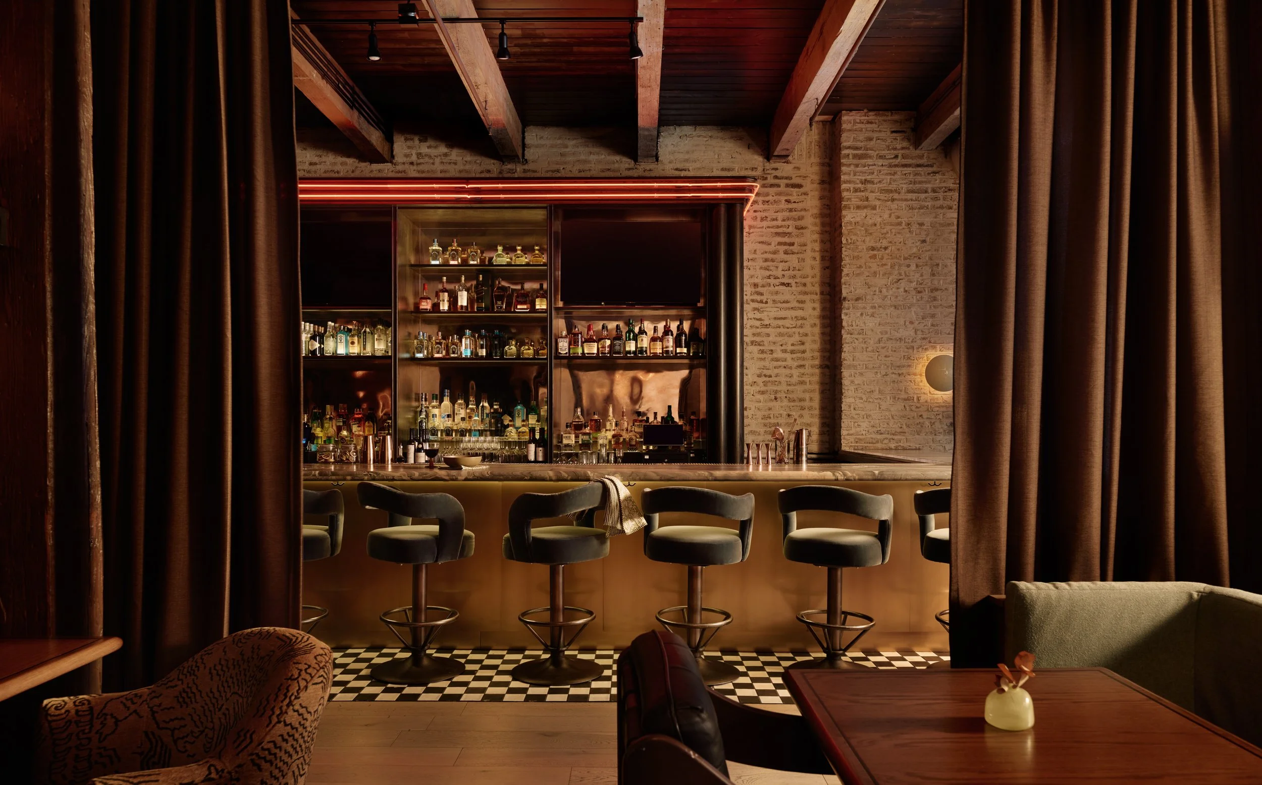 Interior of a modern bar with a long marble counter, barstools, a TV screen showing a basketball game, and shelves with bottles of liquor. Warm lighting and wooden ceiling beams create a cozy atmosphere.