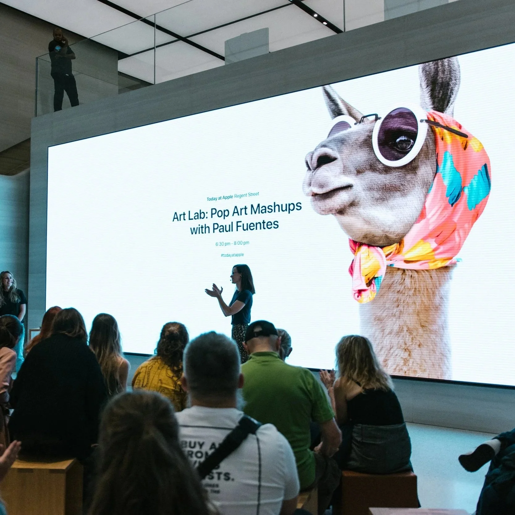 A presenter is speaking to an audience in front of a large screen displaying promotional information for an art event featuring a pop art mashup titled 'Art Lab: Pop Art Mashups with Paul Fuentes'. The screen shows a graphic of a llama wearing sunglasses, a floral headscarf, and a colorful wrap, in a pop art style. The event details are from 6:30 pm to 8:00 pm at Apple Regent Street.