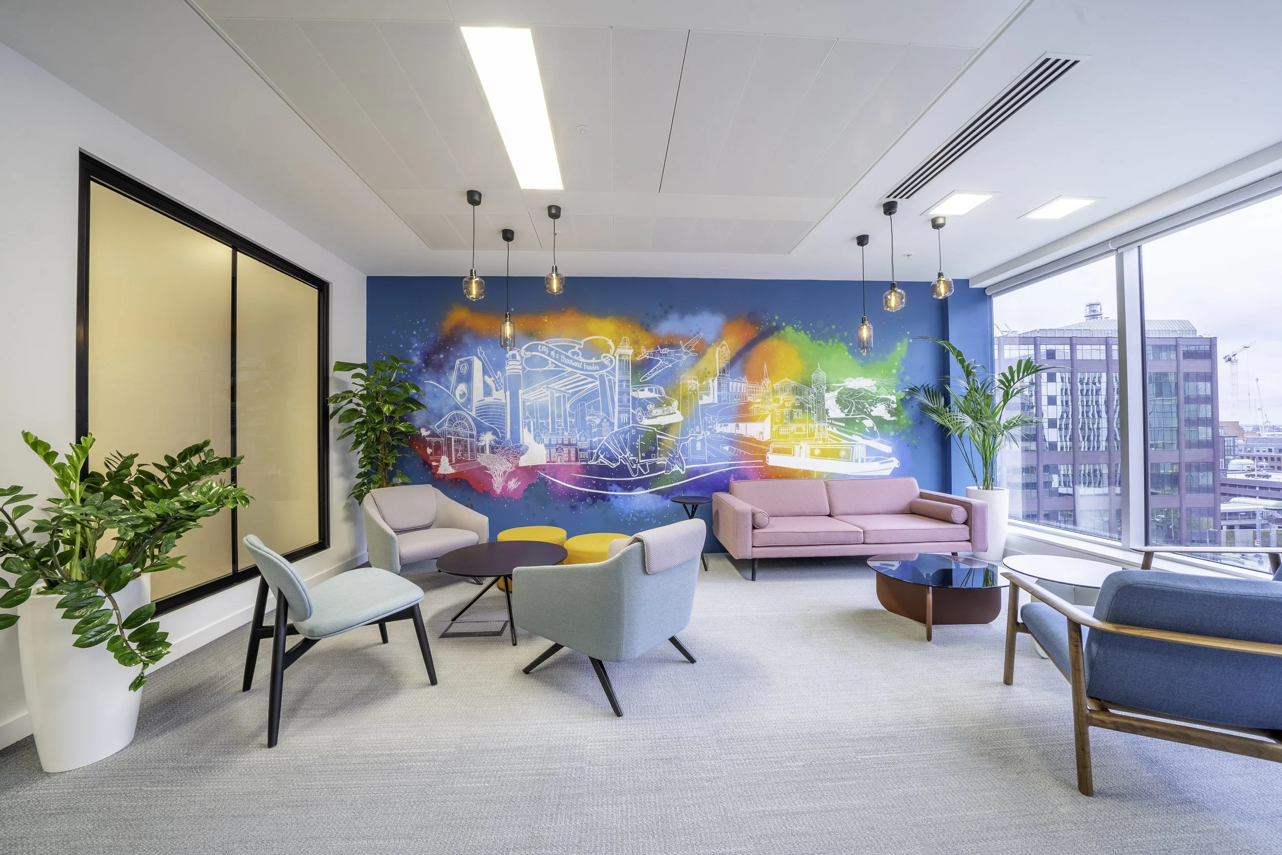 Modern office lounge with colorful mural, pink sofa, blue chairs, and large windows showing city buildings.