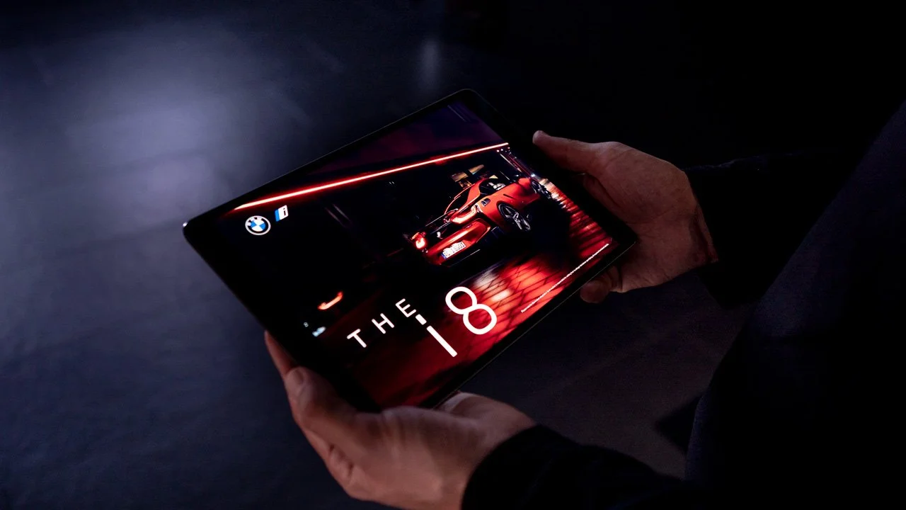 Pessoa segurando um tablet que exibe um anúncio do BMW i8, mostrando um carro vermelho dentro de uma garagem ou showroom escuro.