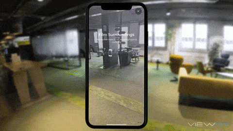 A smartphone displaying an augmented reality view of an office, with the camera turned on and AR markings visible, inside a modern office space with yellow chairs and large windows.