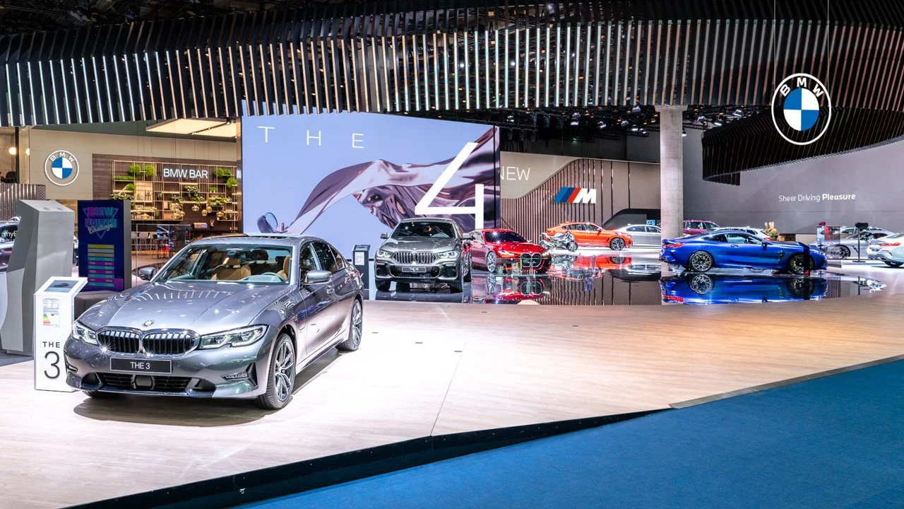 Uma exibição de salão do automóvel com modelos BMW e BMW M, incluindo um BMW Série 3 prateado em primeiro plano e outros carros BMW em várias cores atrás dele, com logotipos da BMW e sinalização promocional.