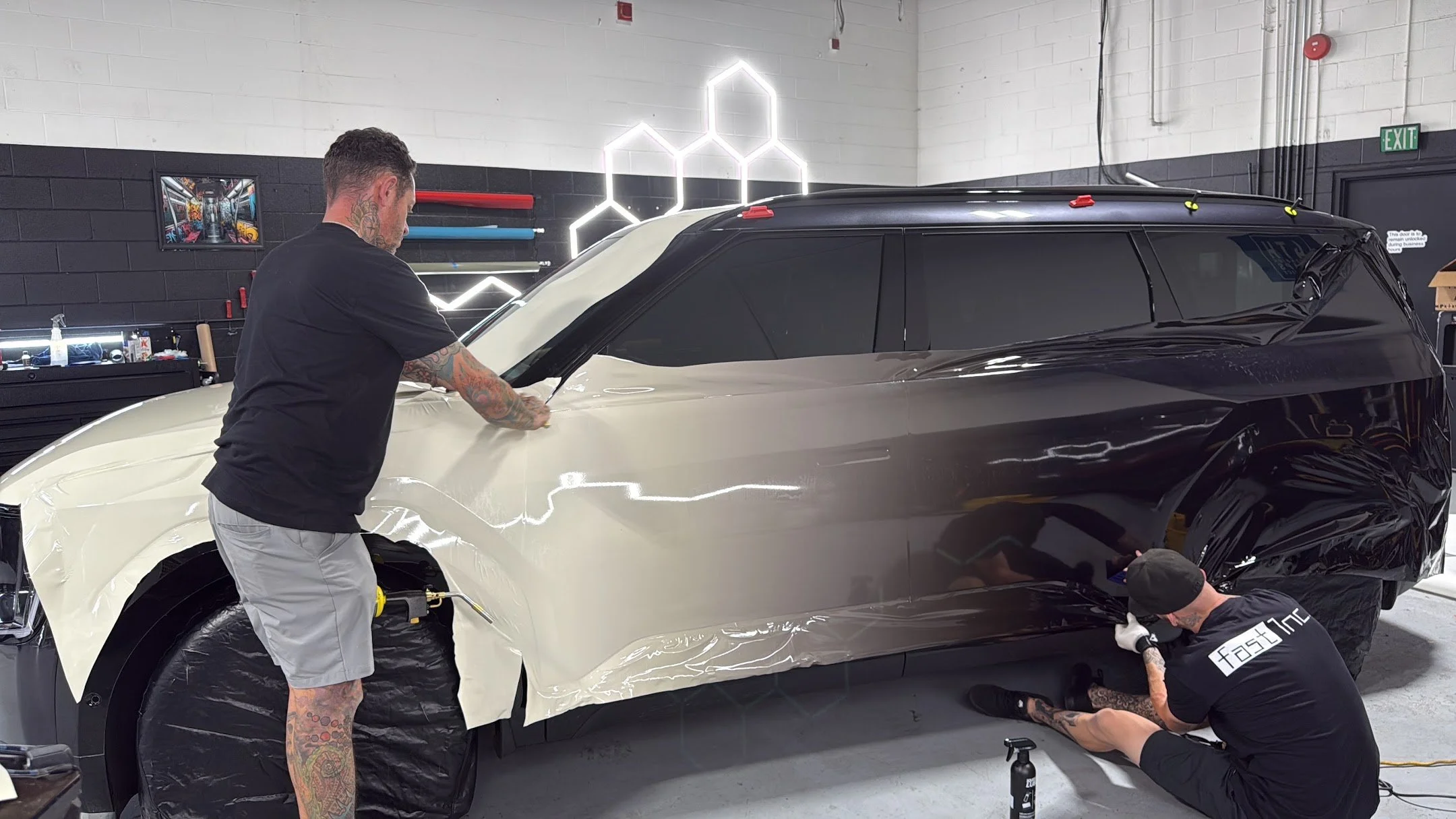 Two workers applying a protective film to a black car in a garage or workshop. The car is partially covered with black and clear film, and the workers are using squeegees and tools to smooth it out.