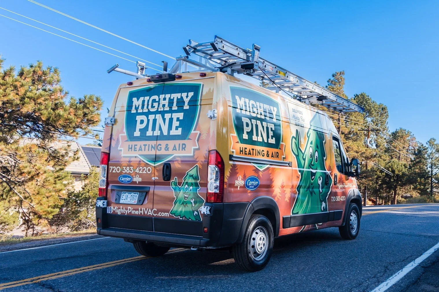 A service van for Mighty Pine Heating & Air with colorful branding and cartoon pine tree mascot, parked on a scenic road with trees and clear blue sky in the background.