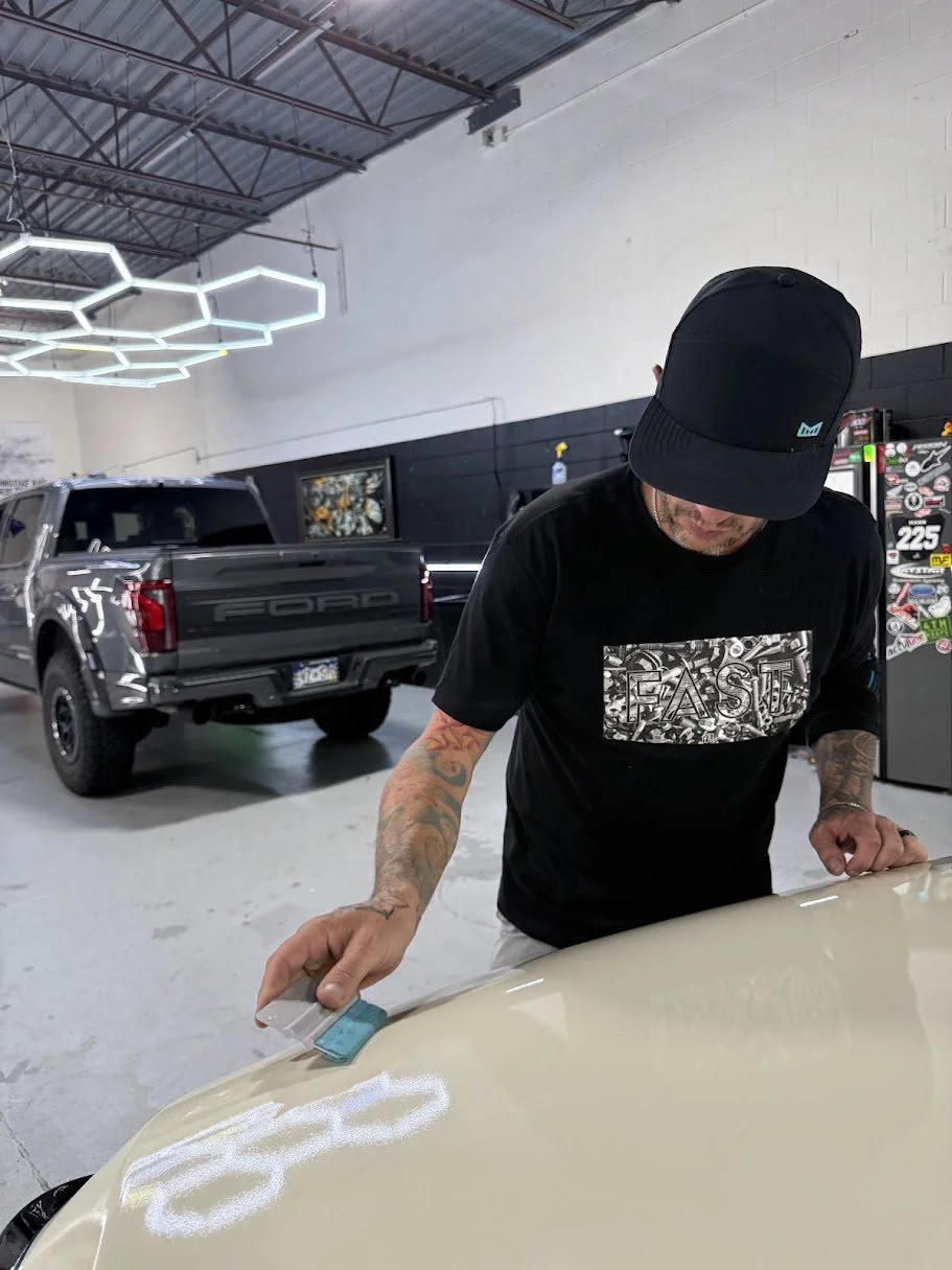 A man with tattoos on his arms, wearing a black cap and black T-shirt, is working on a beige car in an indoor garage. There is a gray Ford truck parked in the background and modern hexagon-shaped ceiling lights.