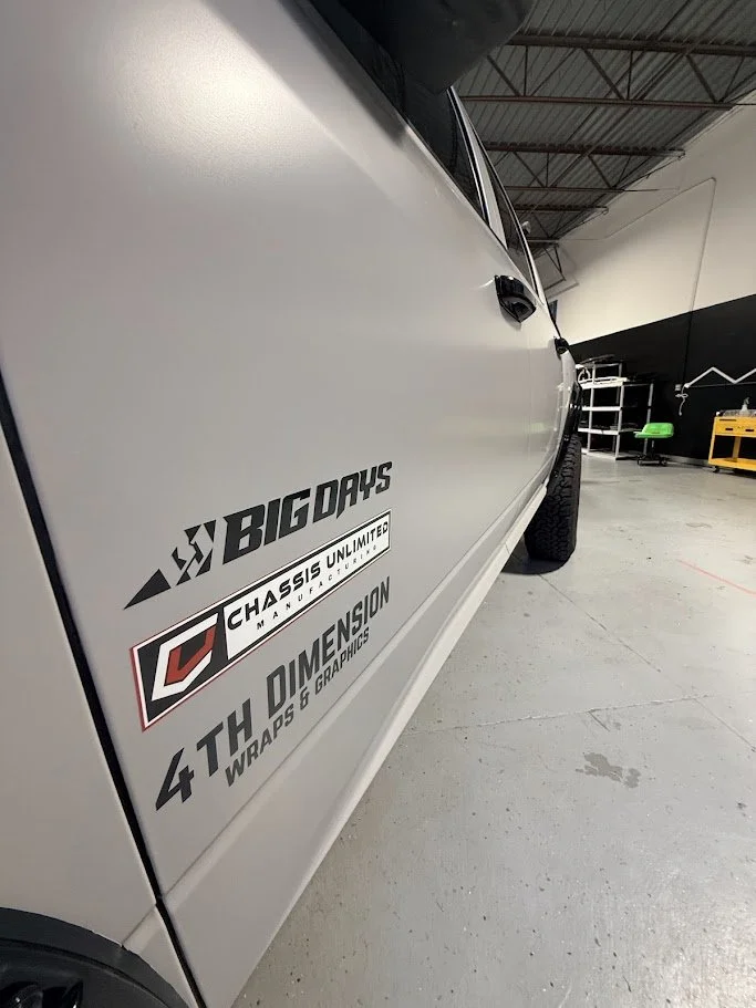 Close-up of the side of a white vehicle with black lettering logos for BIG DAYS, CHASSIS UNLIMITED, and 4TH DIMENSION WRAPS & GRAPHICS, in a garage or workshop.