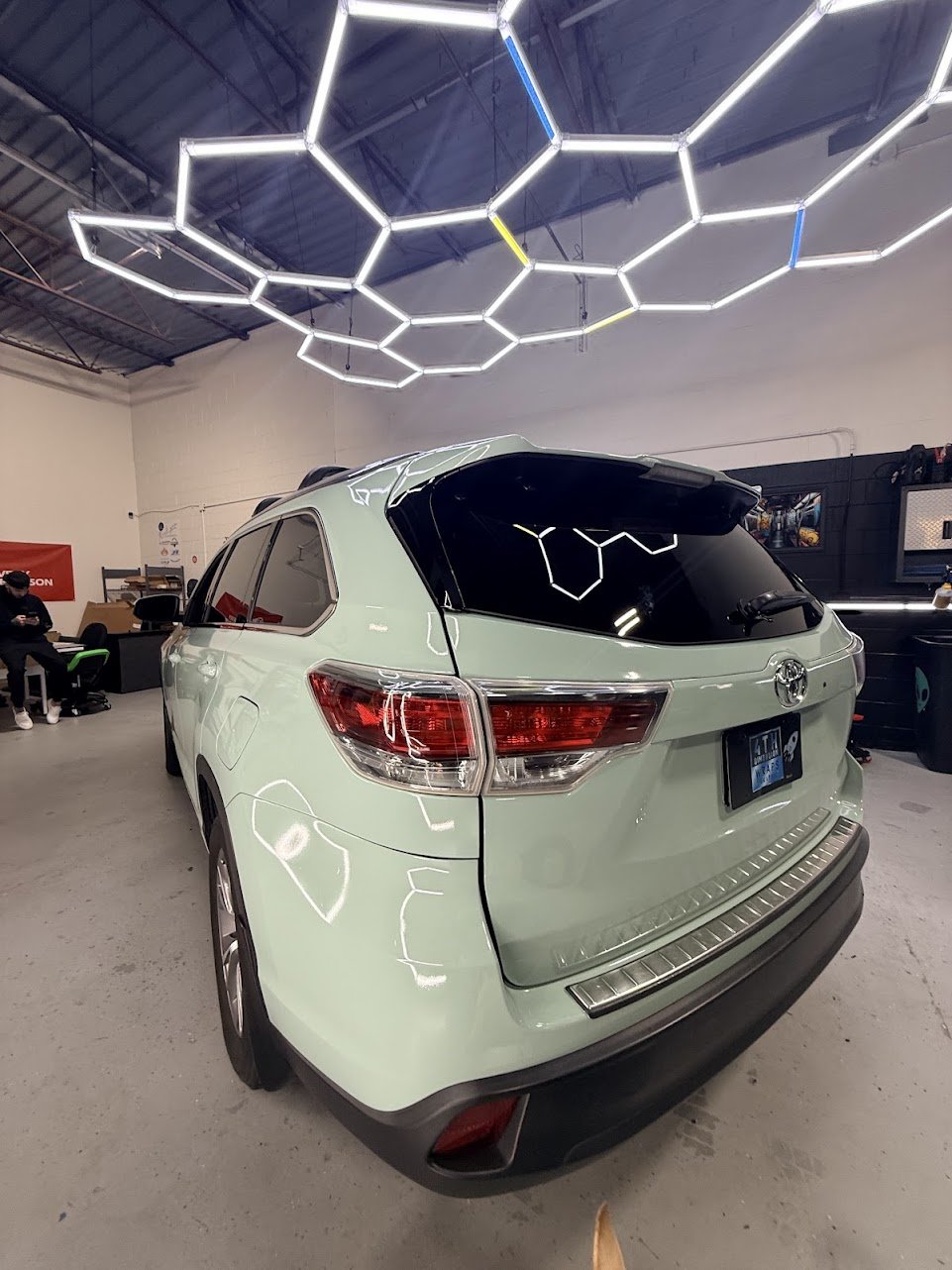 Light green Toyota SUV in an indoor space with hexagonal LED light fixtures on the ceiling and some equipment and seating area in the background.