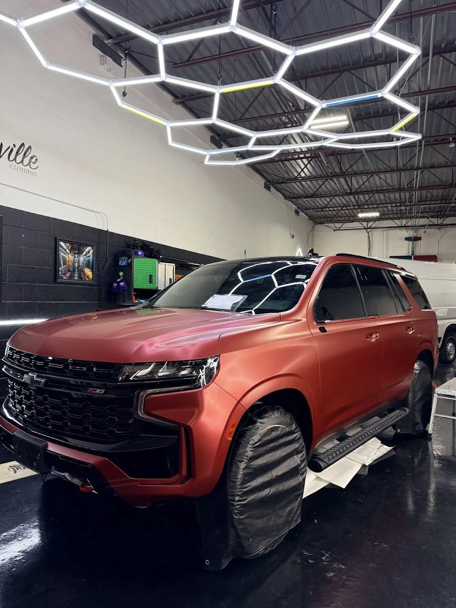 Red SUV parked indoors with car cover on tires, modern lighting fixture overhead, and a white wall in the background.