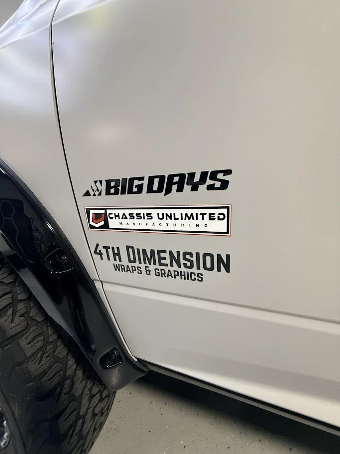 Close-up of a white vehicle showing stickers that read 'BIG DAYS,' 'Chassis Unlimited Manufacturing,' and '4th Dimension Wraps & Graphics' near the wheel.