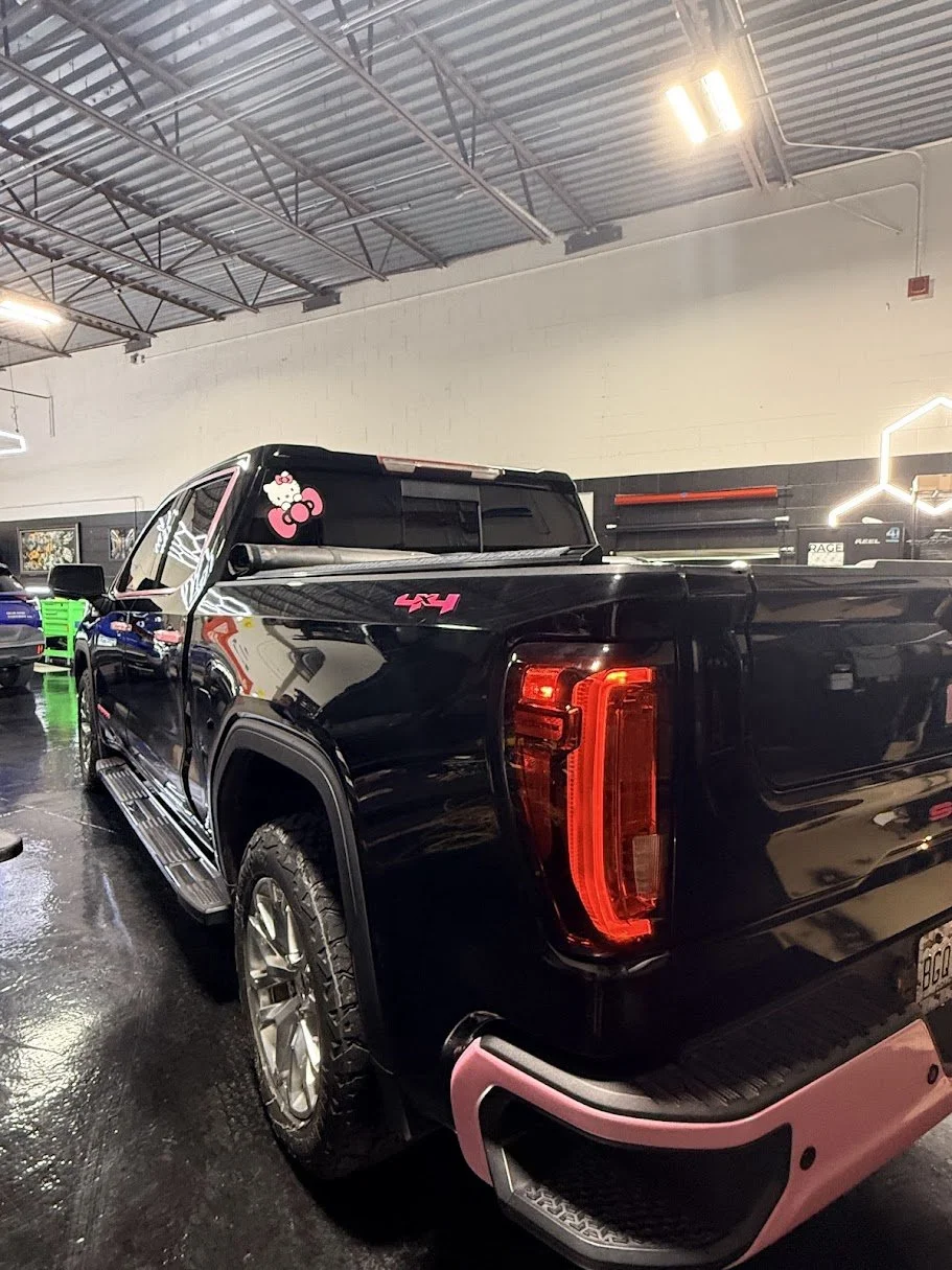 Black pickup truck with pink accents, Hello Kitty sticker on the window, parked inside a showroom or garage with black and white walls and overhead lighting.