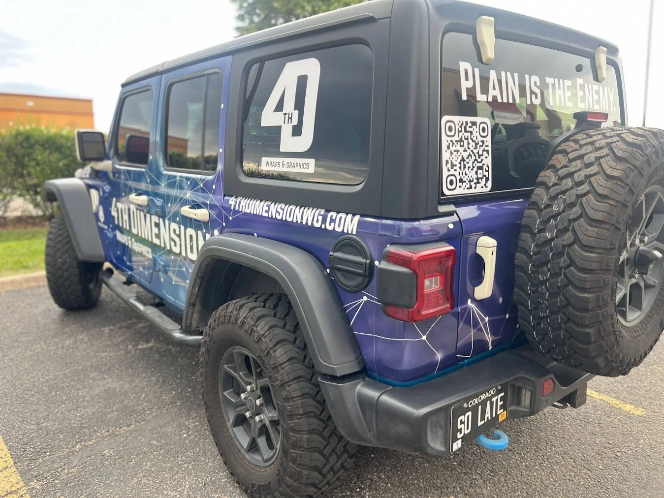 A customized black and blue Jeep wrapped in graphics for 4th Dimension Wraps & Graphics. The vehicle has large off-road tires, a spare tire on the back, and a humorous Colorado license plate that reads "SO LATE." The back window displays a QR code, a