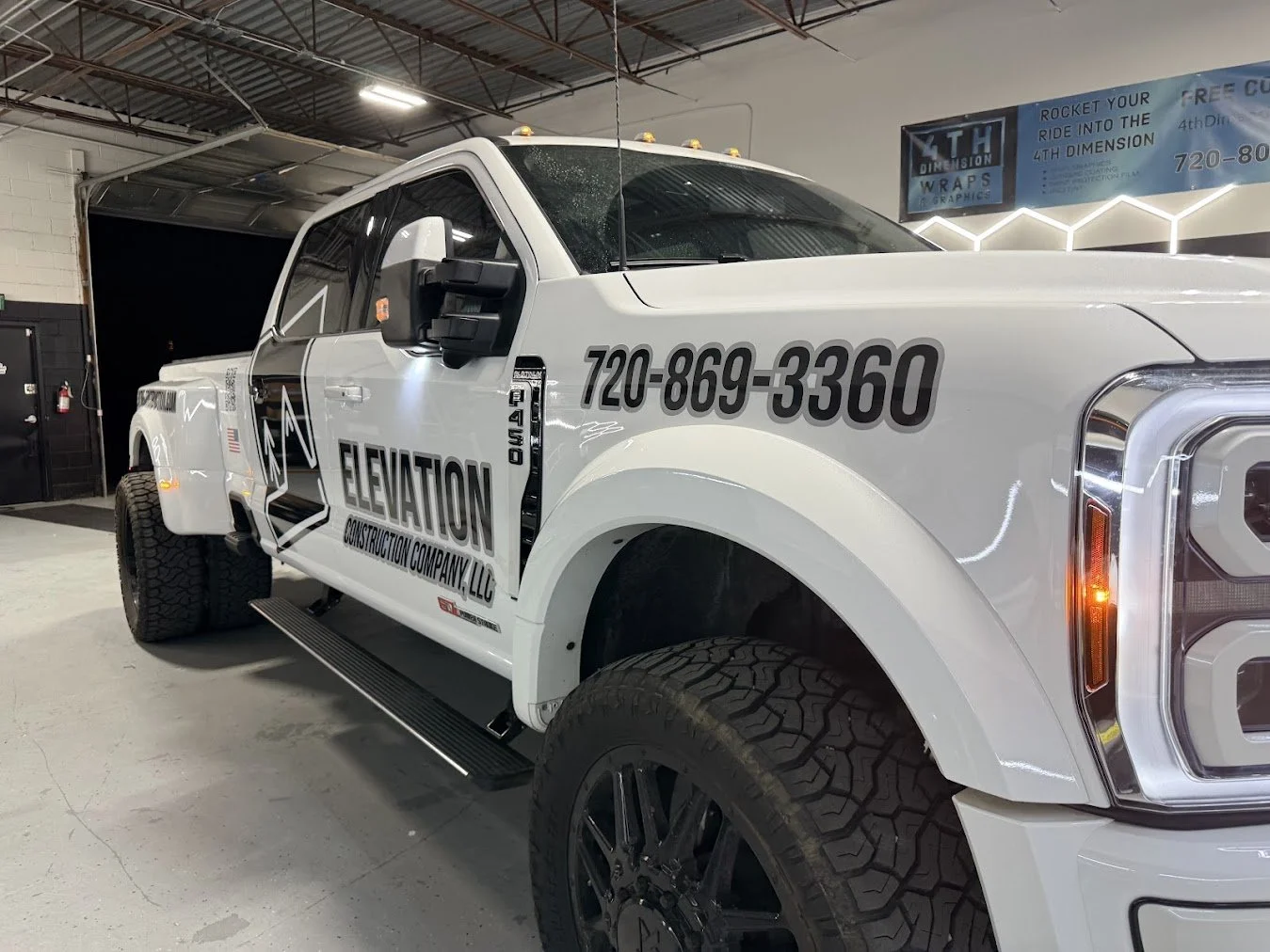 A white pickup truck with the logo and contact information for Elevation Construction Company, LLC, is parked inside a garage or showroom. The truck has large off-road tires and black decals on the side.