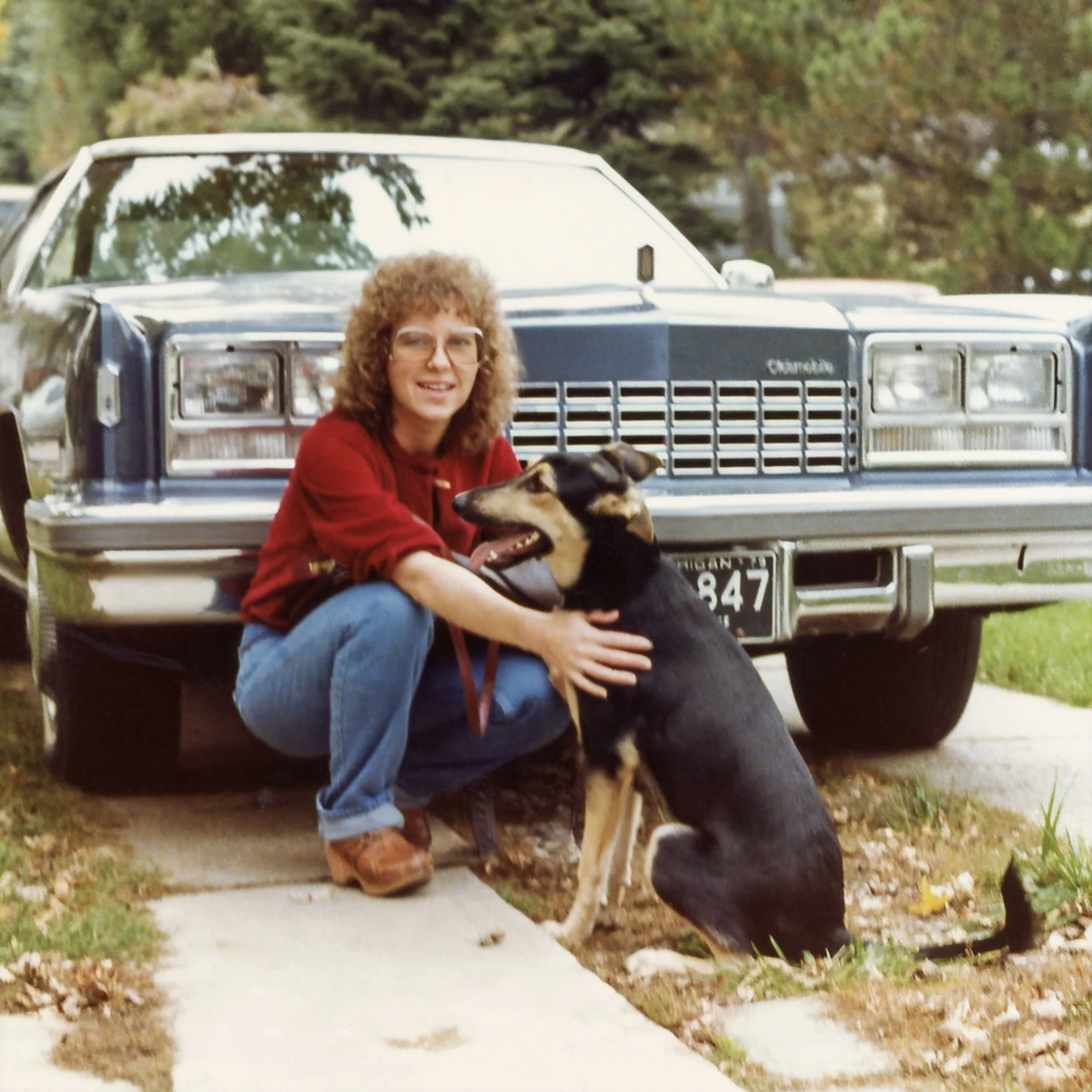Roni is kneeling with Mugsy in front of her 1977 land yacht, an Oldsmobile Toronado. 