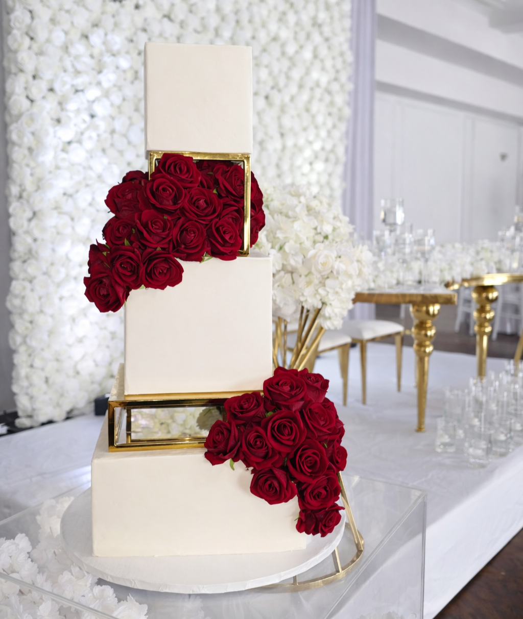 A wedding cake decorated with red roses and gold accents, displayed in an elegant reception hall with white floral walls and gold table decor.