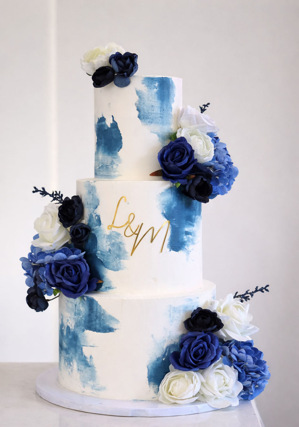 Three-tiered wedding cake decorated with blue, white, and black roses and blue watercolor-style frosting, with a gold cursive name "Leah" on the middle tier.