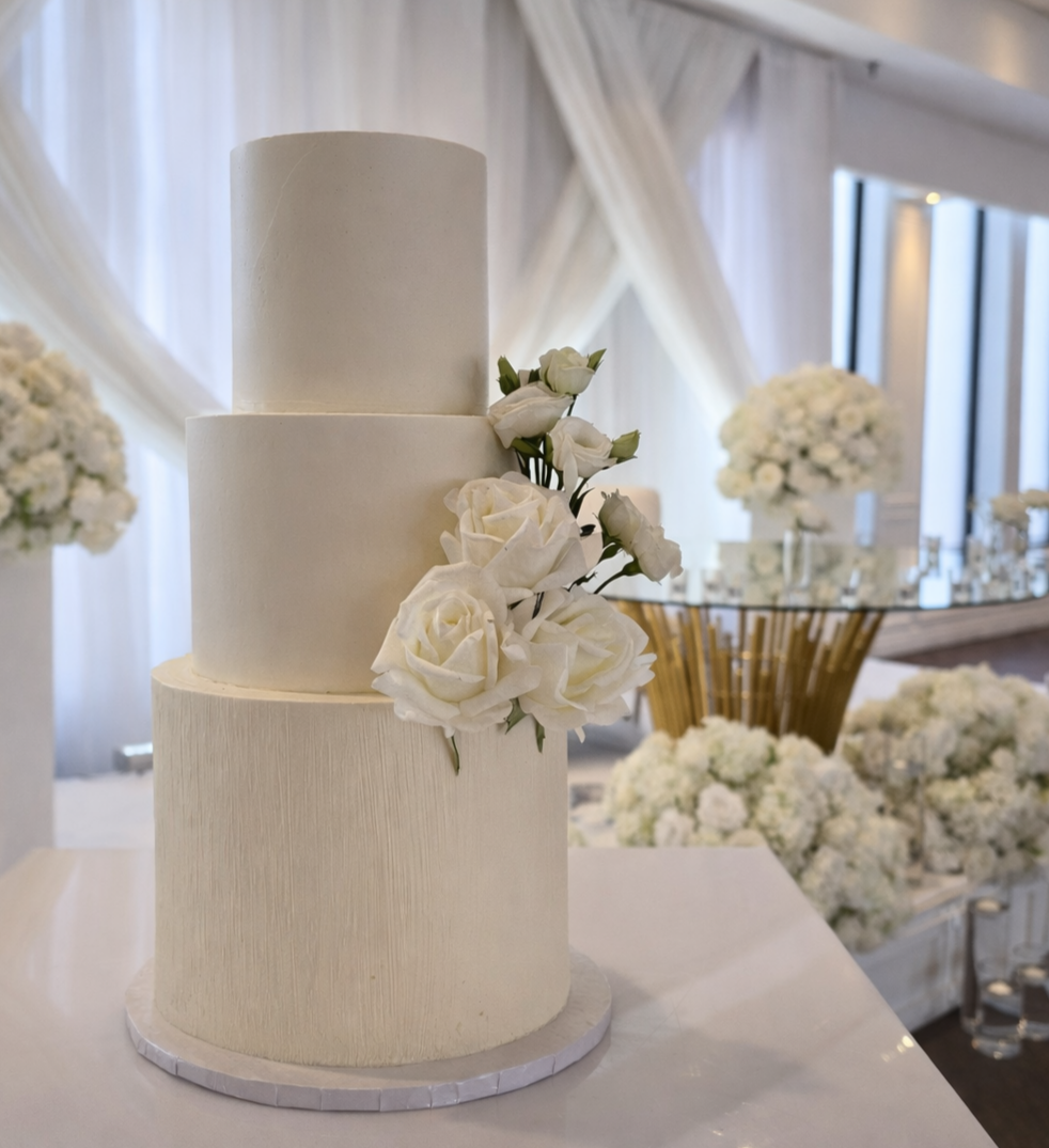 Three-tier white wedding cake with a cluster of white roses and greenery, arranged on a white table with a floral backdrop and a gold and glass stand in the background.