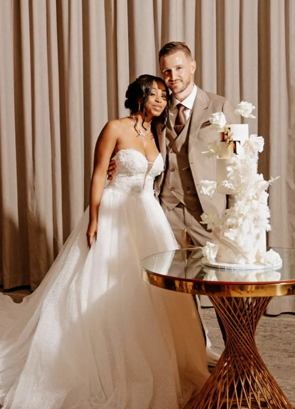 A bride and groom at their wedding reception, standing together beside a tiered white wedding cake with gold accents and decorative flowers.