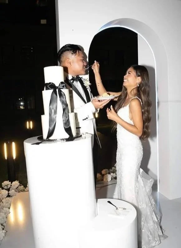 Bride and groom celebrating at their wedding, feeding each other cake, with a tall white wedding cake decorated with black bow. 