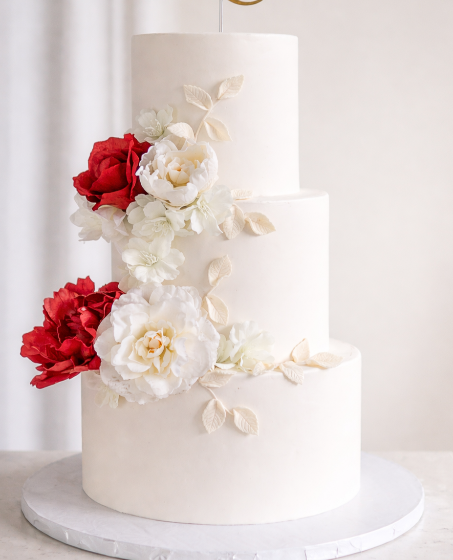 Three-tier white wedding cake decorated with white and red flowers and leaves, on a white cake board.