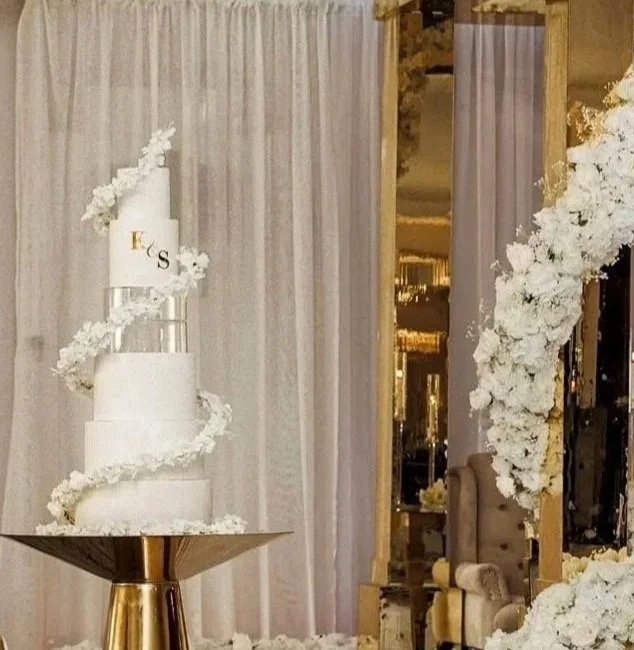 A four-tier white wedding cake with cascading white flowers, placed on a gold stand in front of white curtains, with floral arrangements and a mirror in the background.