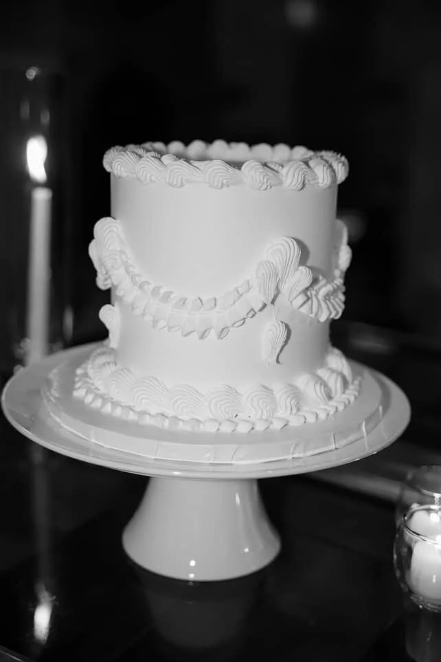 A white, decorated birthday or celebration cake with ornate piped icing patterns on a display stand.