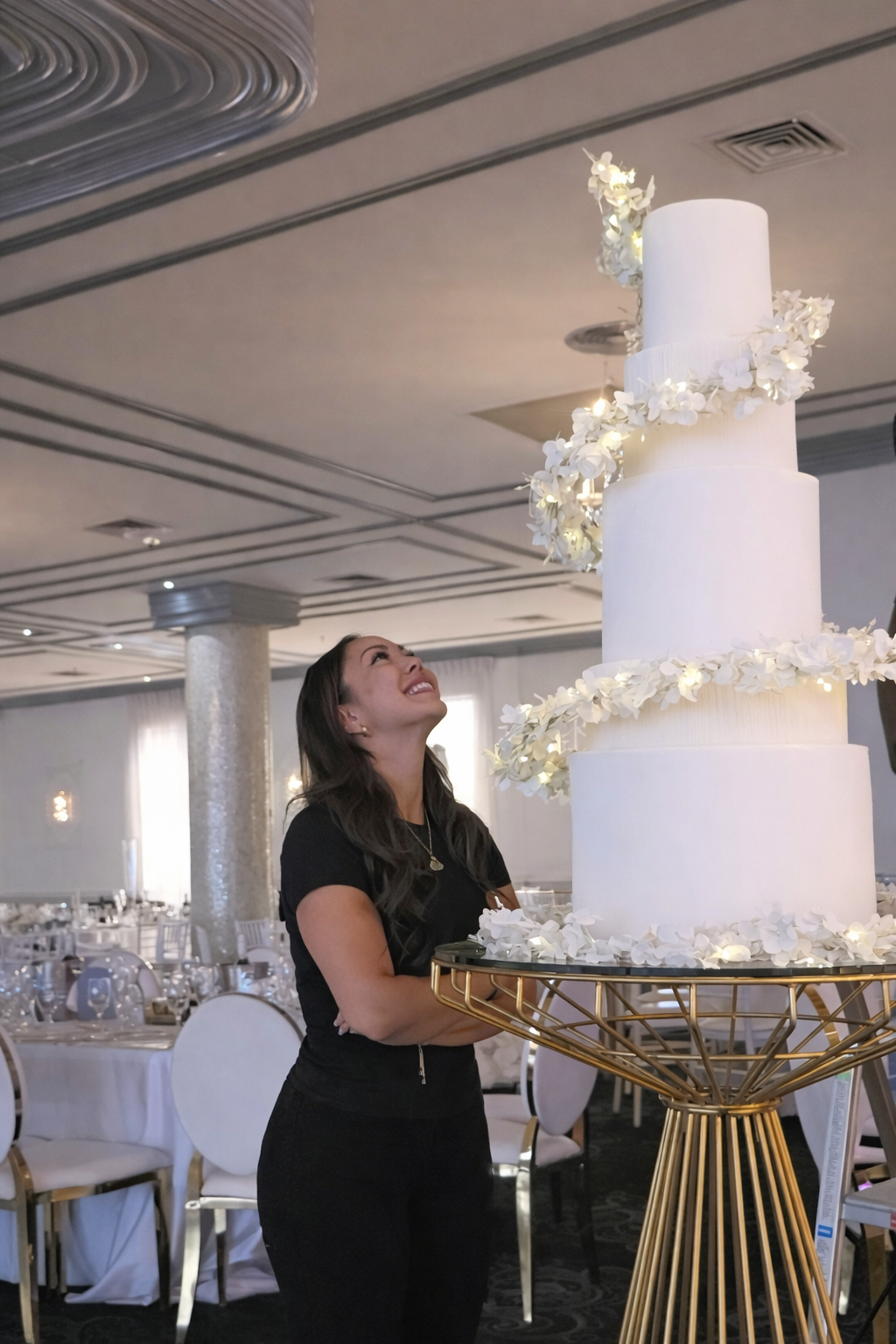 Woman in black dress smiling at a tall white wedding cake decorated with white flowers in an elegant reception hall.