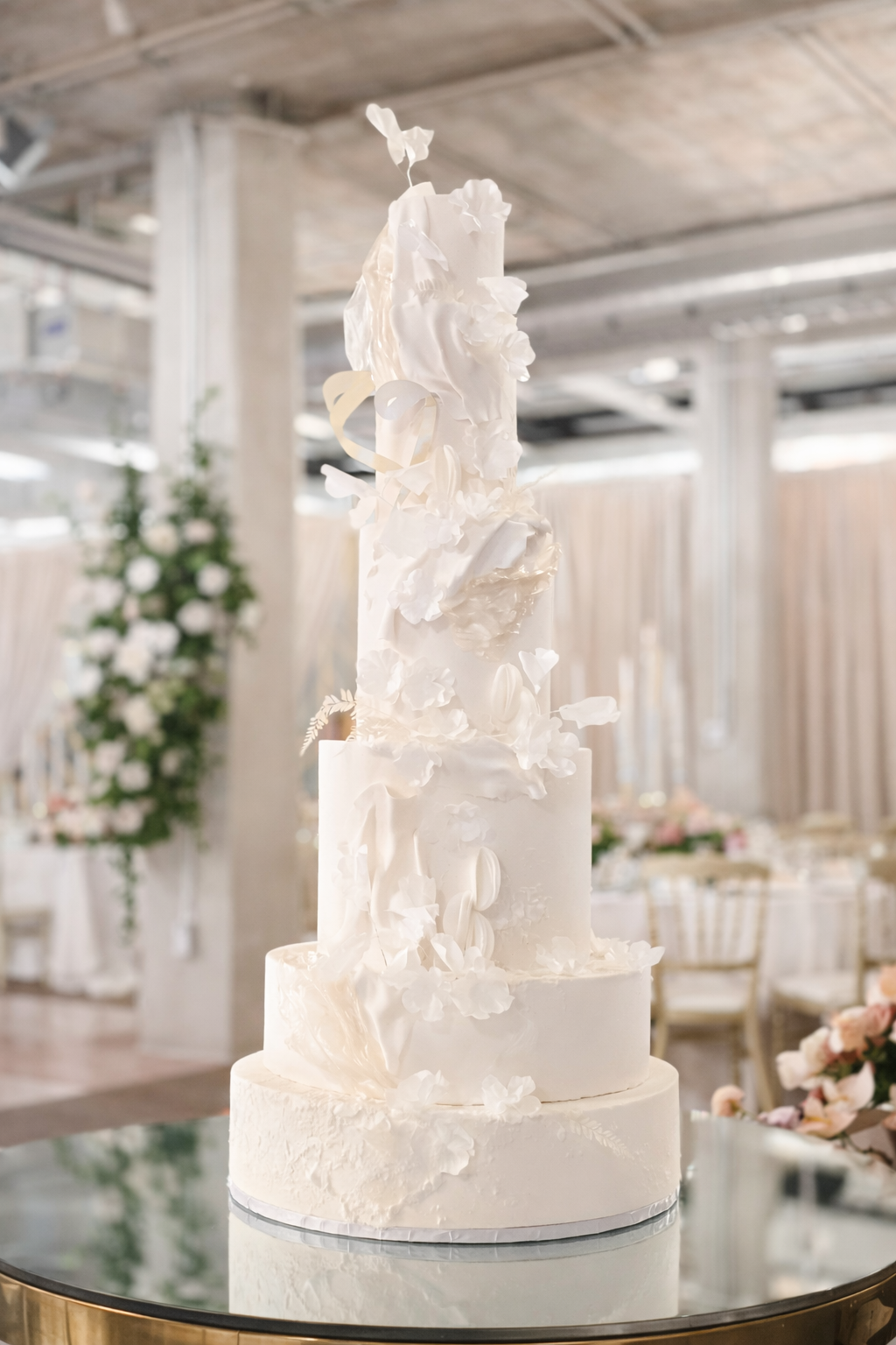 A tall, four-tiered white wedding cake with delicate floral decorations on each layer, displayed on a reflective round table in a decorated event space.
