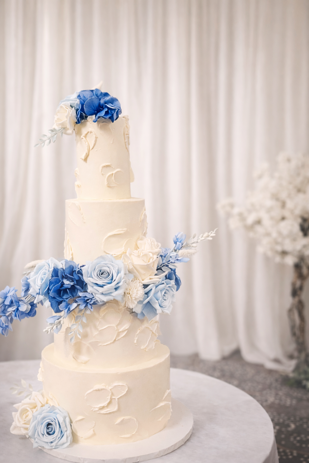 A four-tier white wedding cake decorated with blue and white flowers, set on a white table with a light background.