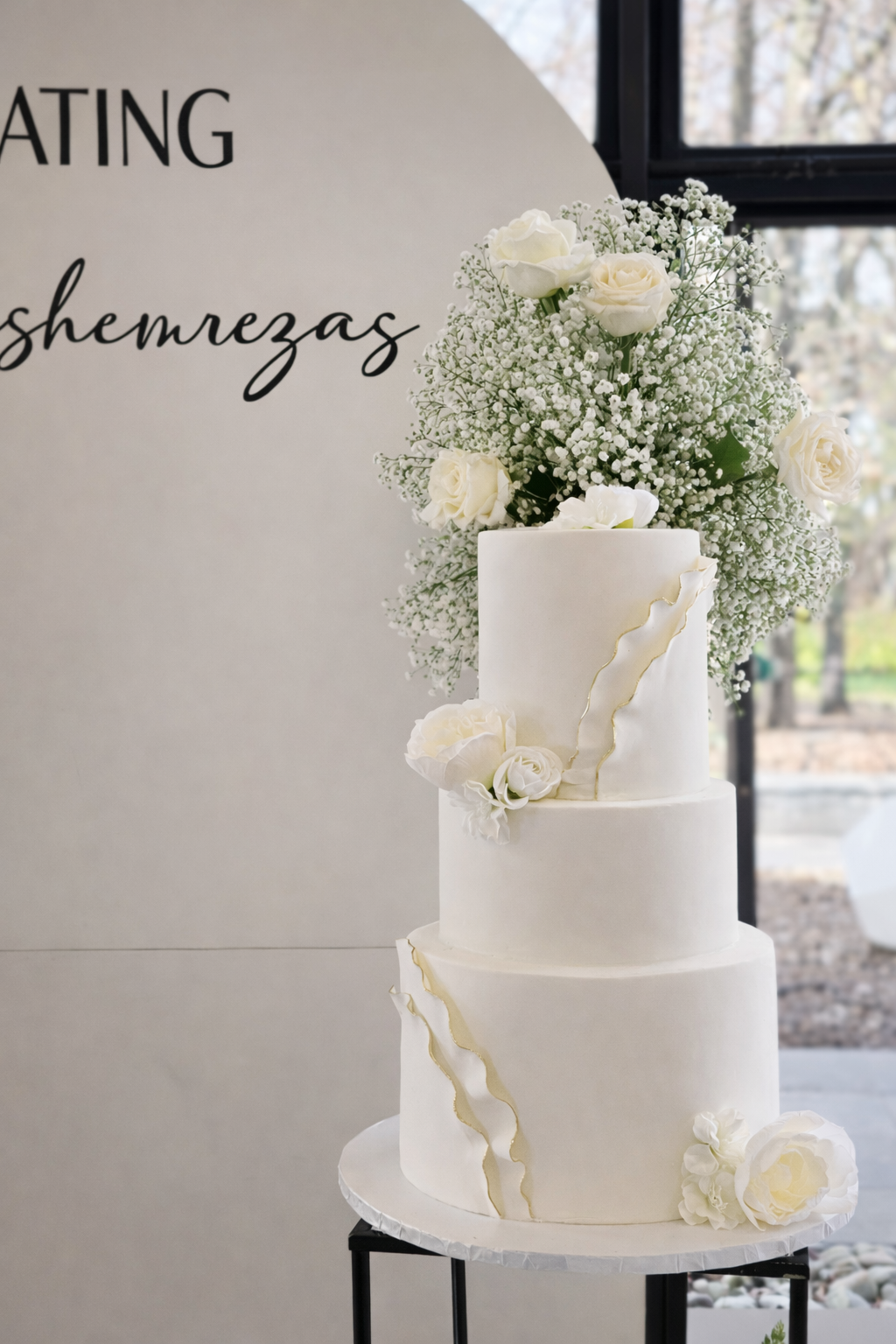 A three-tier white wedding cake decorated with white roses, peonies, and baby's breath, with gold-colored wavy accents, placed on a black stand.