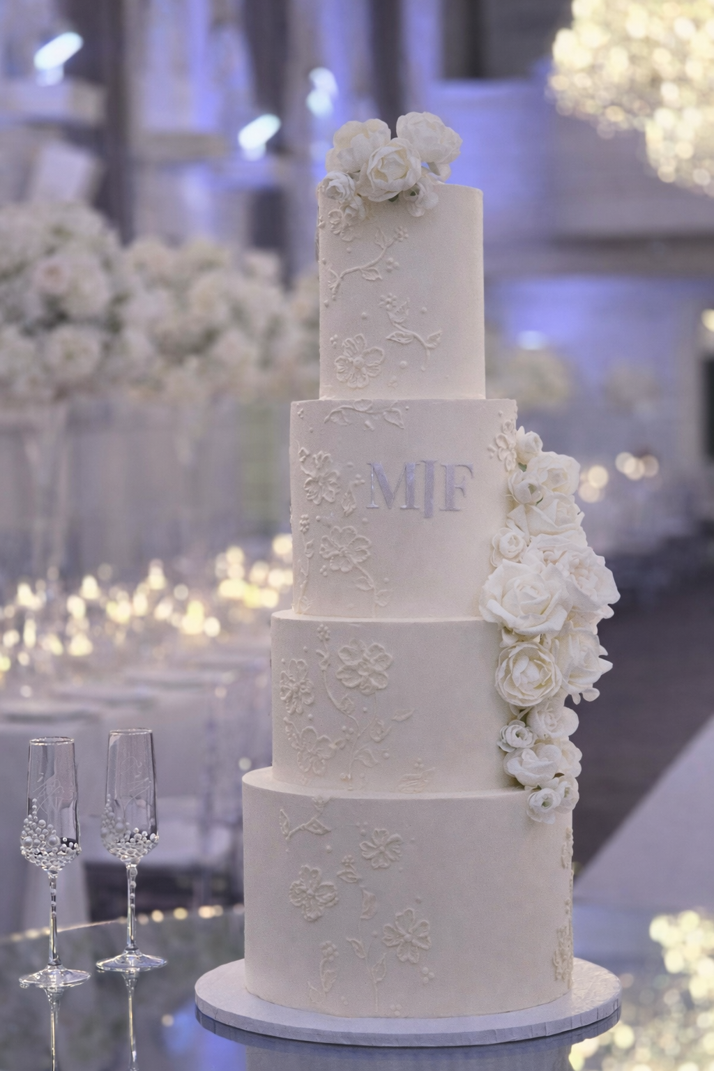 A tall, four-tier white wedding cake decorated with intricate floral patterns and white roses. The middle tier has the initials 'MIF' in silver. The cake is set on a round white stand with champagne glasses beside it, and a blurred background with fl